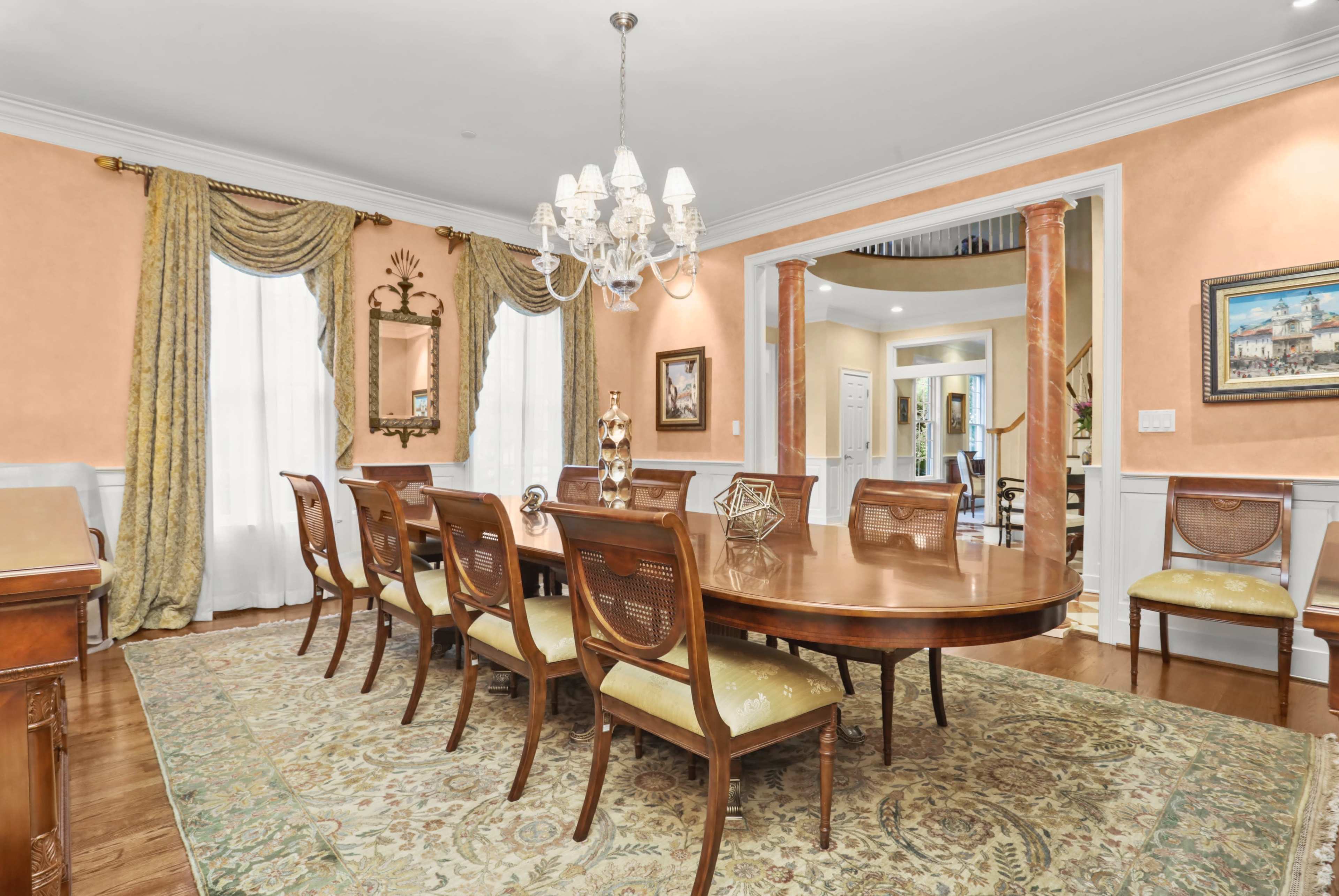 A spacious dining room with a large wooden table surrounded by twelve chairs, enhanced by an elegant chandelier and decorative curtains.