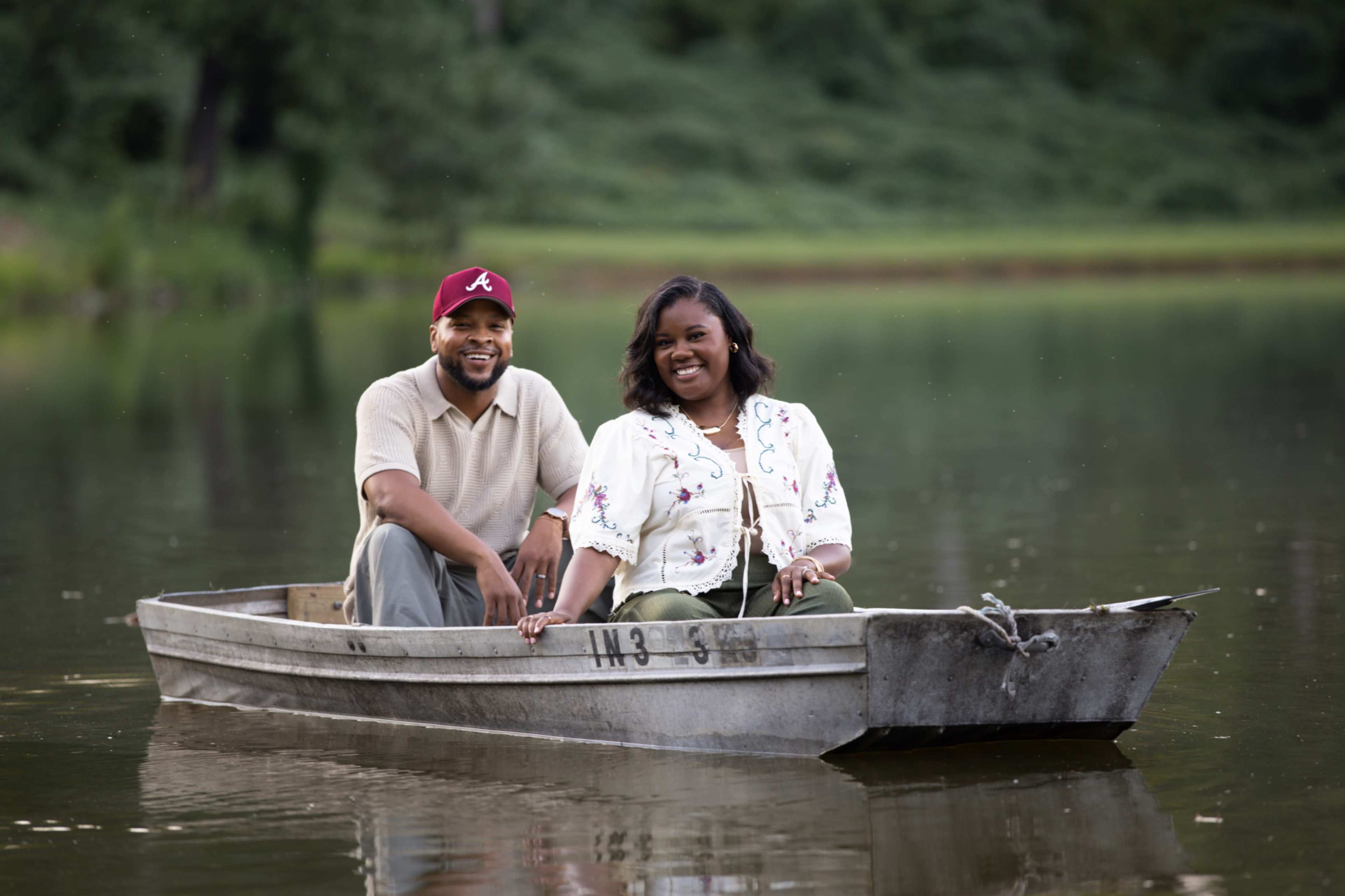 A man and a woman sit together in a small boat on a calm lake surrounded by greenery.