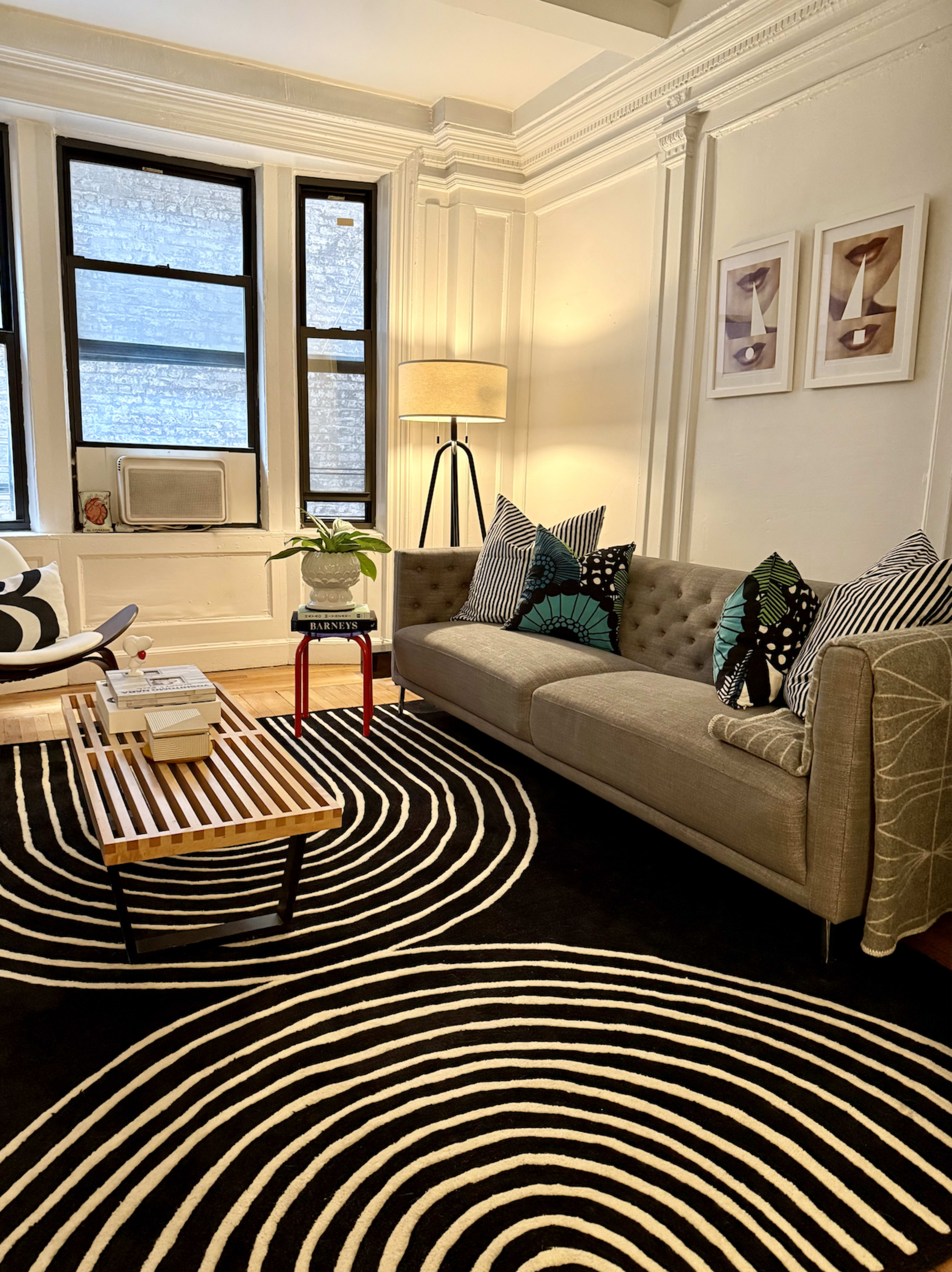 A modern living room features a gray couch adorned with patterned pillows, a striped rug, a wooden coffee table, and an elegant lamp beside large windows.