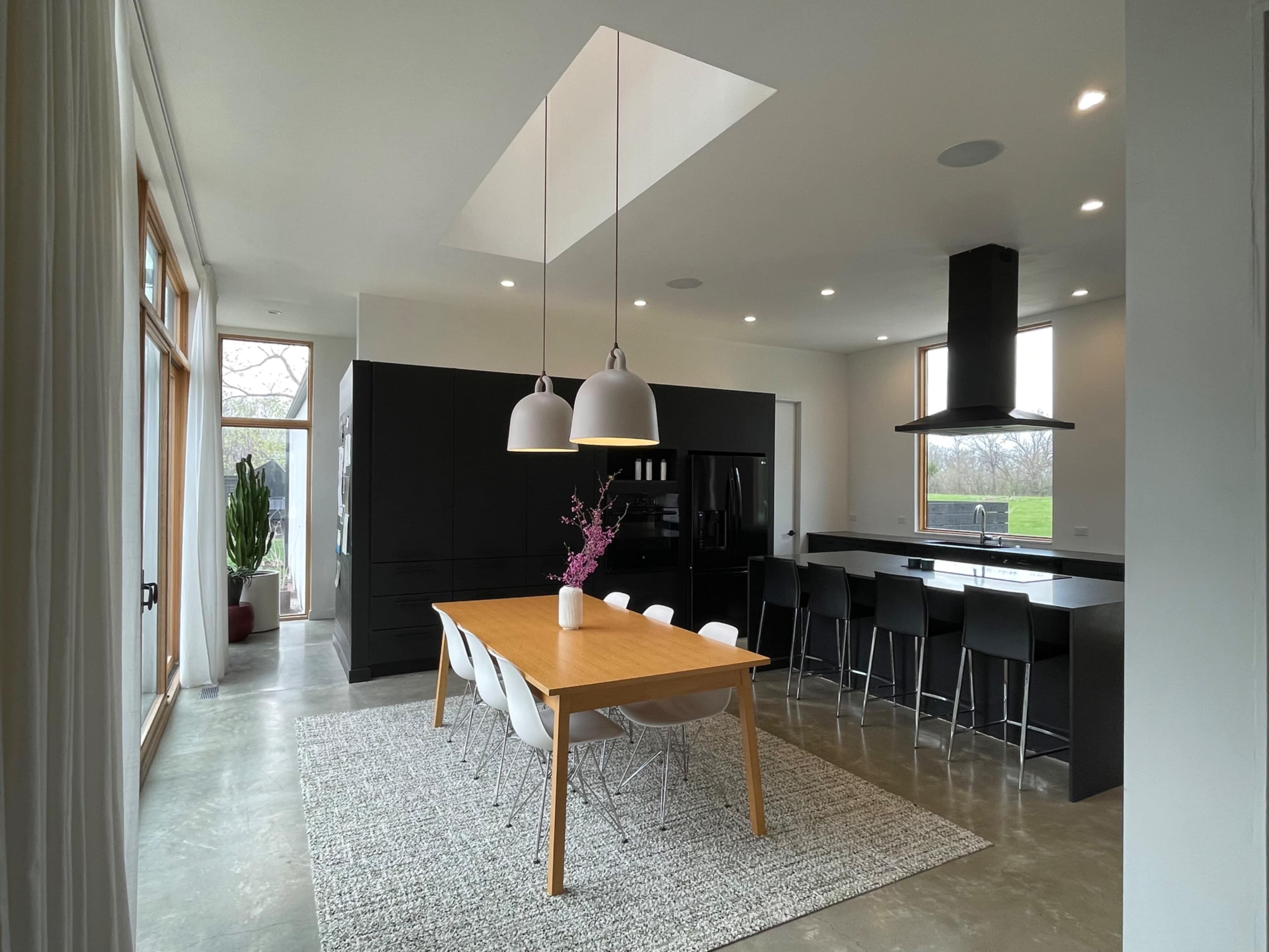 A modern kitchen features a wooden dining table with white chairs, paired with black cabinetry and large windows.