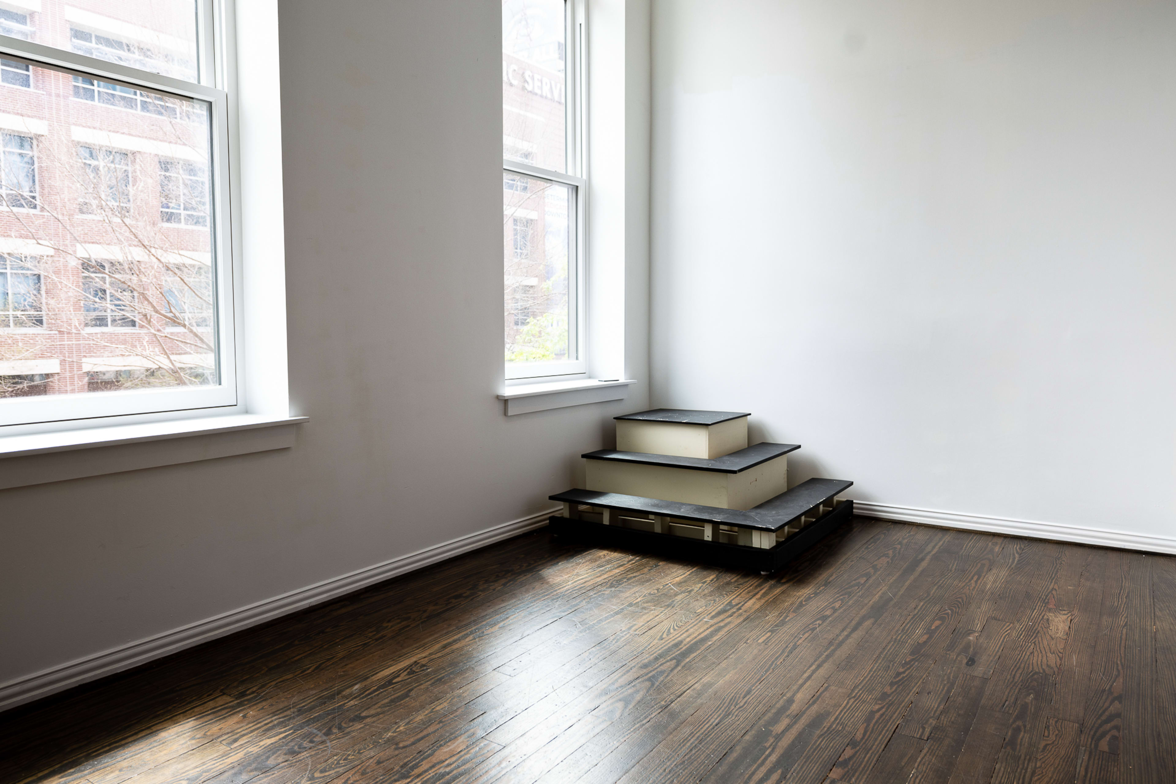 A corner of a room features large windows beside a tiered wooden platform on the floor.