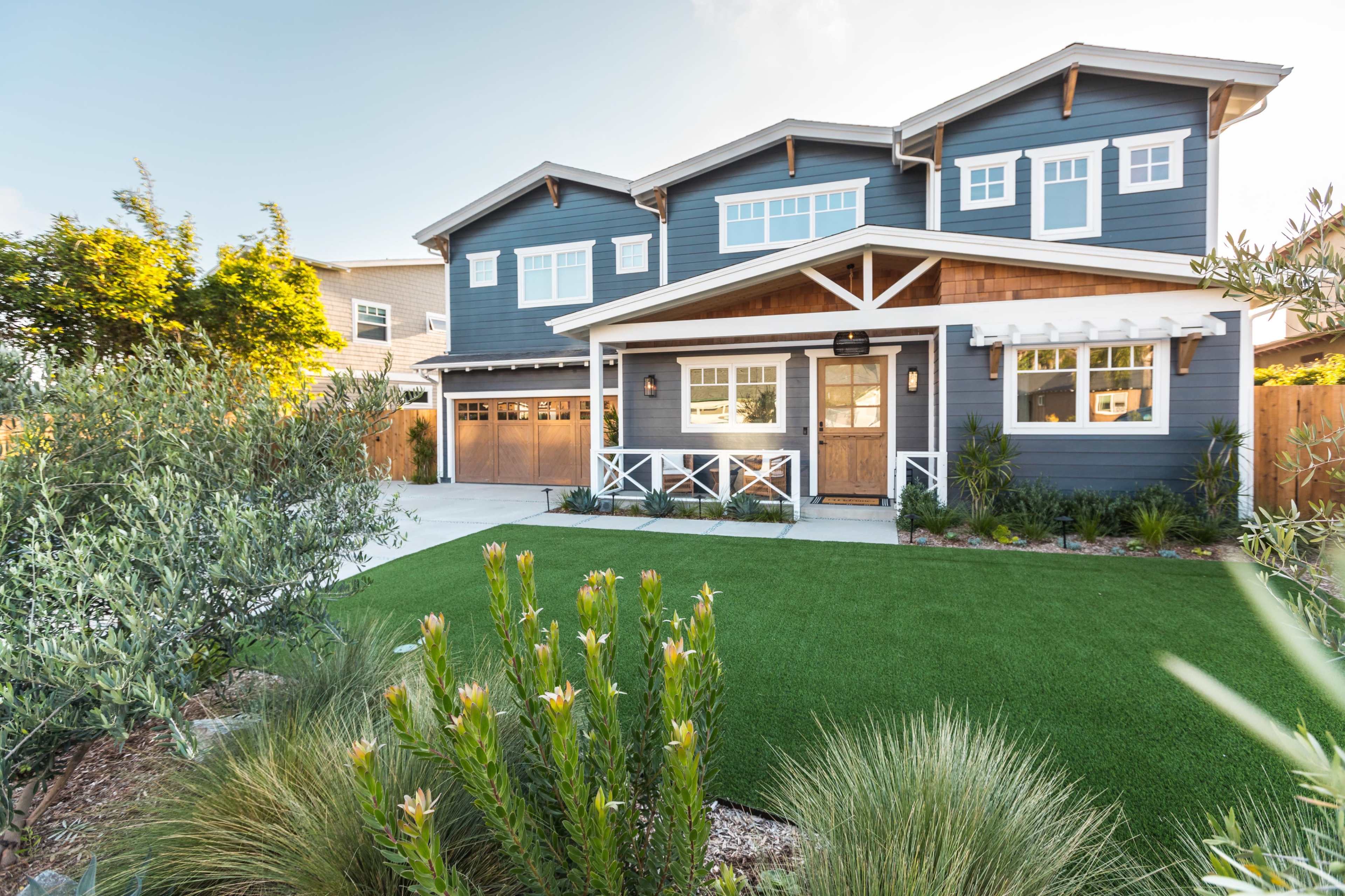 A two-story blue house with a wooden porch and garage doors is surrounded by landscaped greenery and artificial turf.