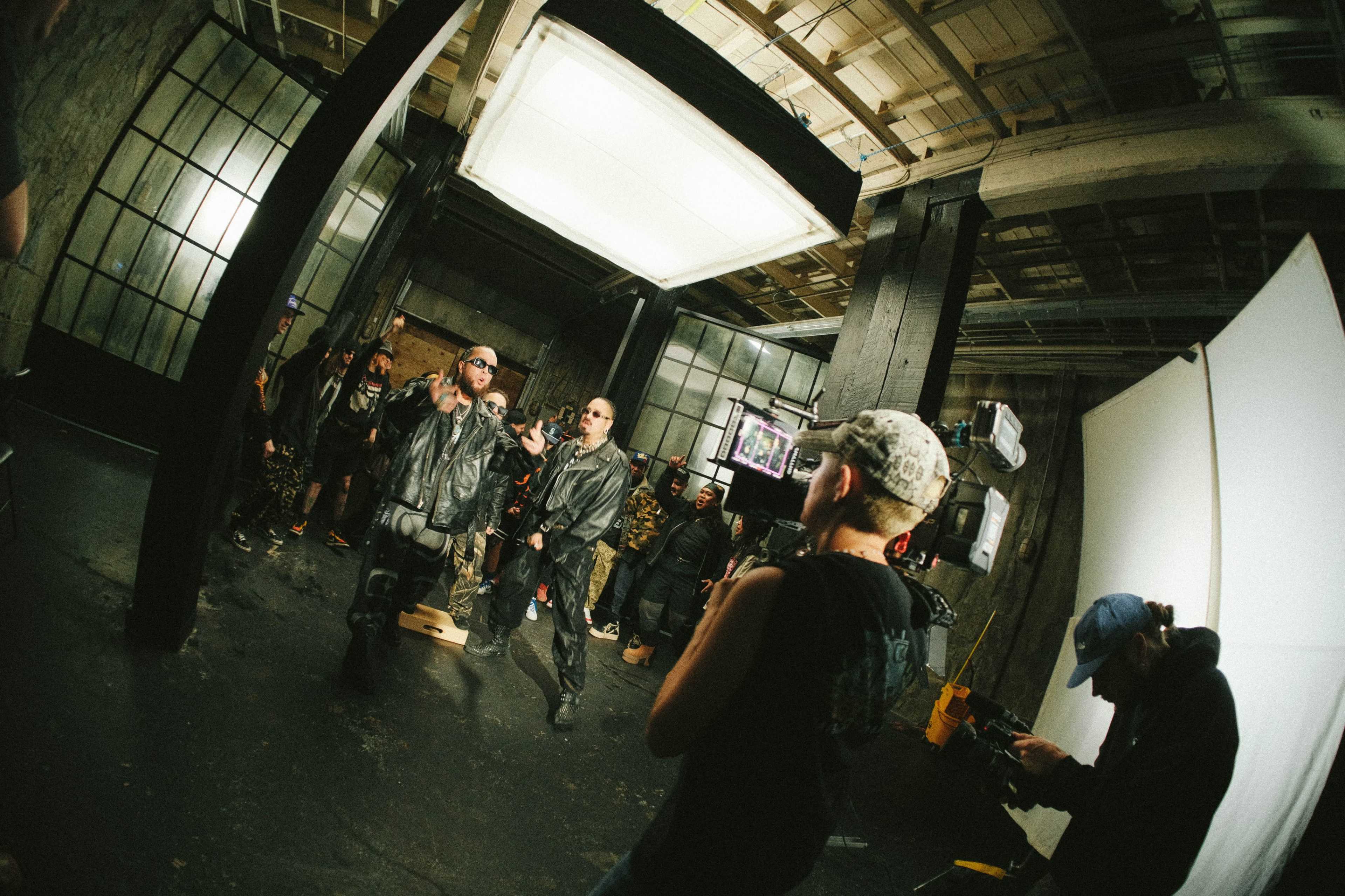 The image shows a group of people in a dimly lit studio, with some wearing leather jackets and sunglasses, and a crew member adjusting equipment in the foreground.