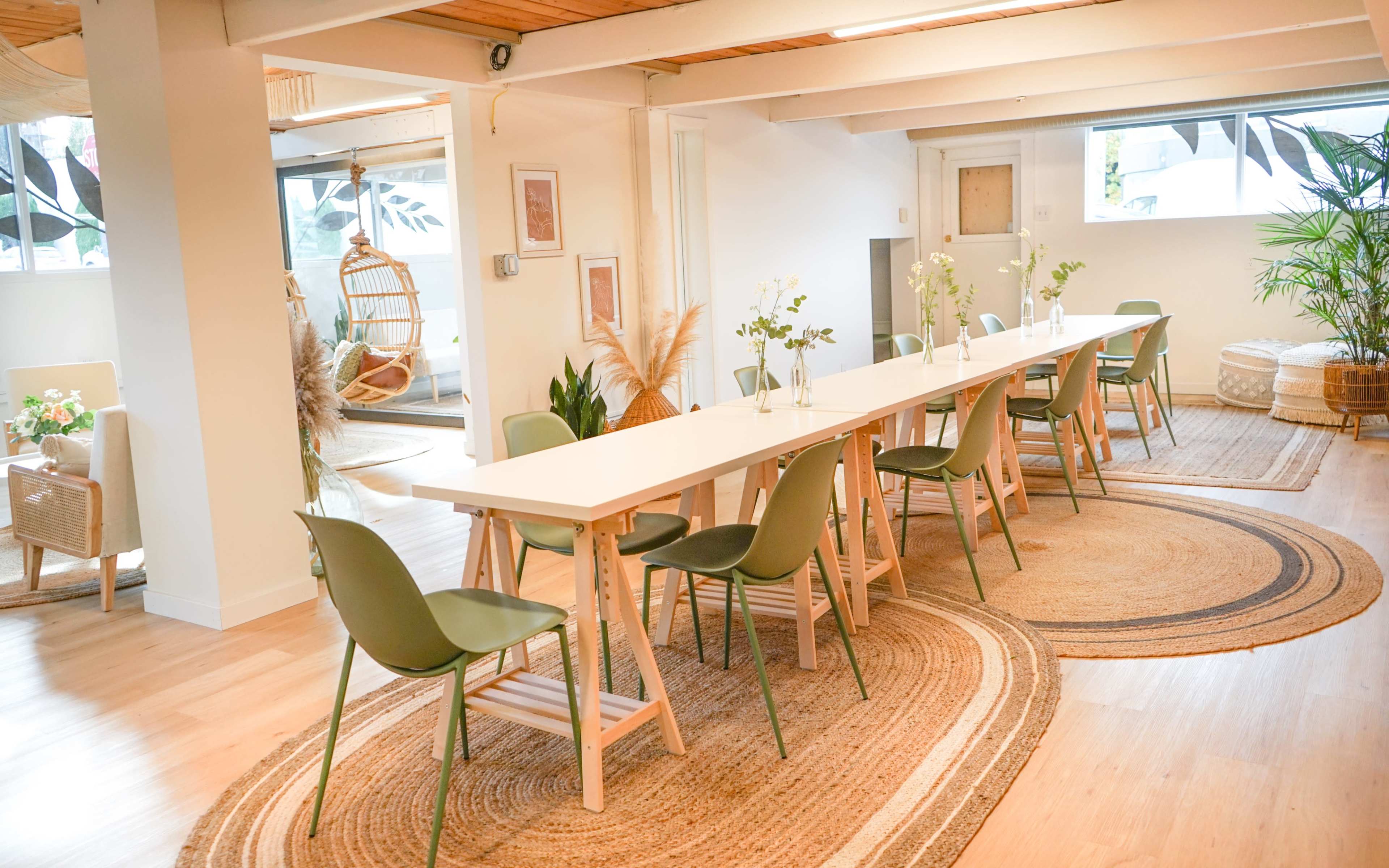 A spacious room features a long white table surrounded by green chairs, with decorative plants and woven rugs enhancing the cozy atmosphere.