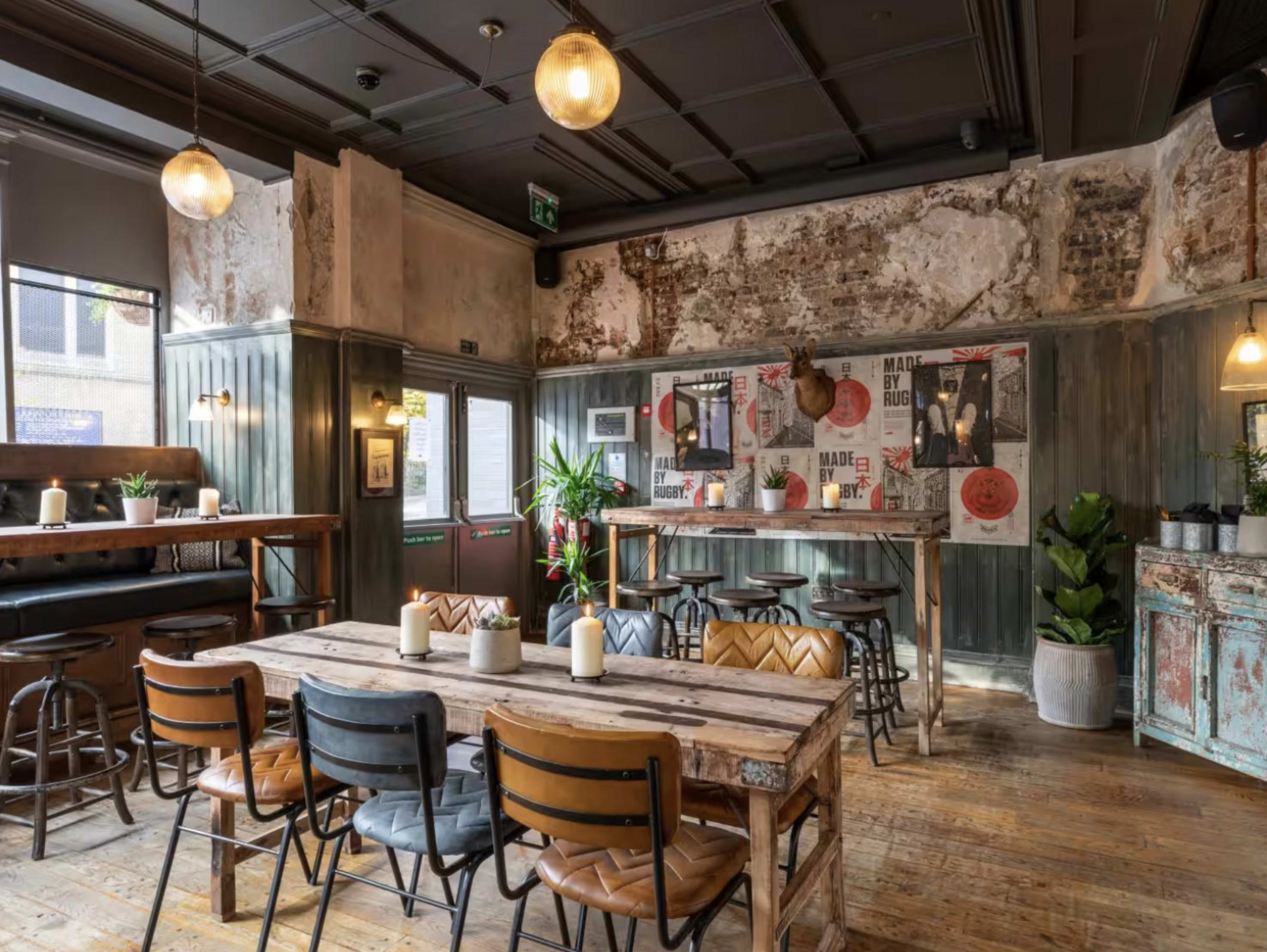 The image shows an interior of a rustic cafe or bar with wooden seating, a communal table, and vintage decor featuring plants and wall art.