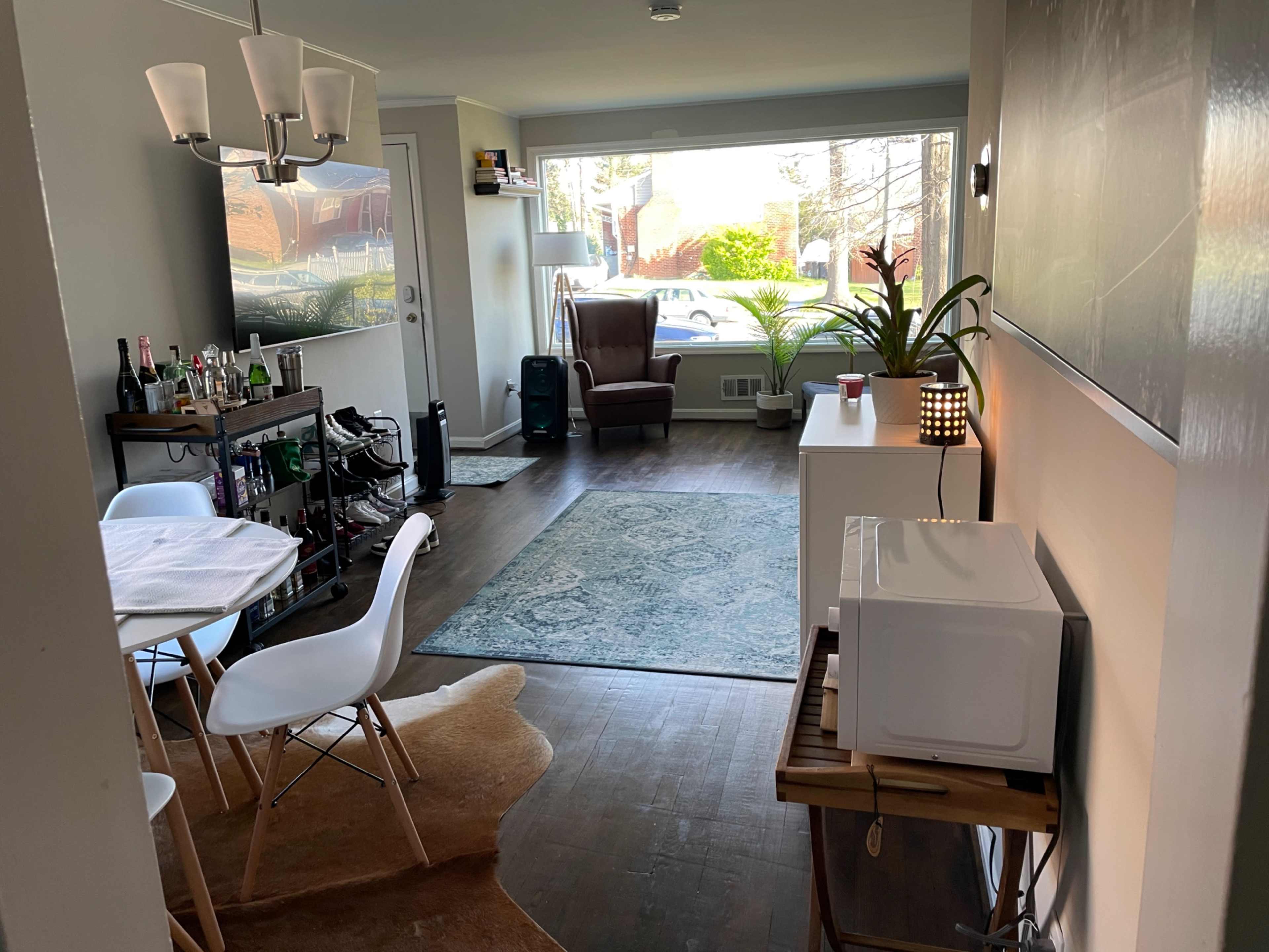 A hallway leads to a living area with a dining table, a large window, and a comfortable chair, featuring a plant and a microwave.
