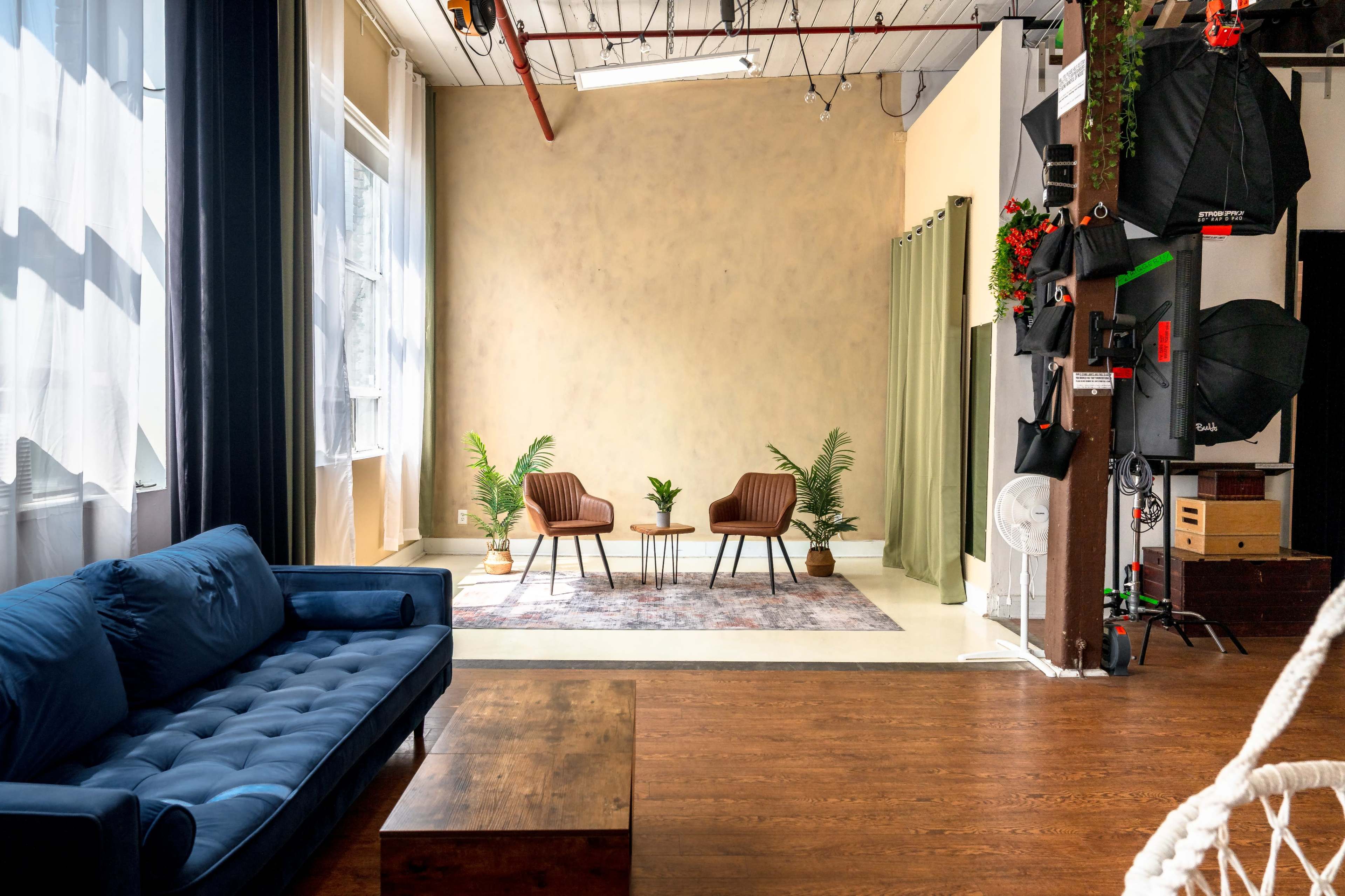 The image shows a spacious interior with a blue sofa, a wooden coffee table, two brown chairs, and a plant in front of a light-colored wall.