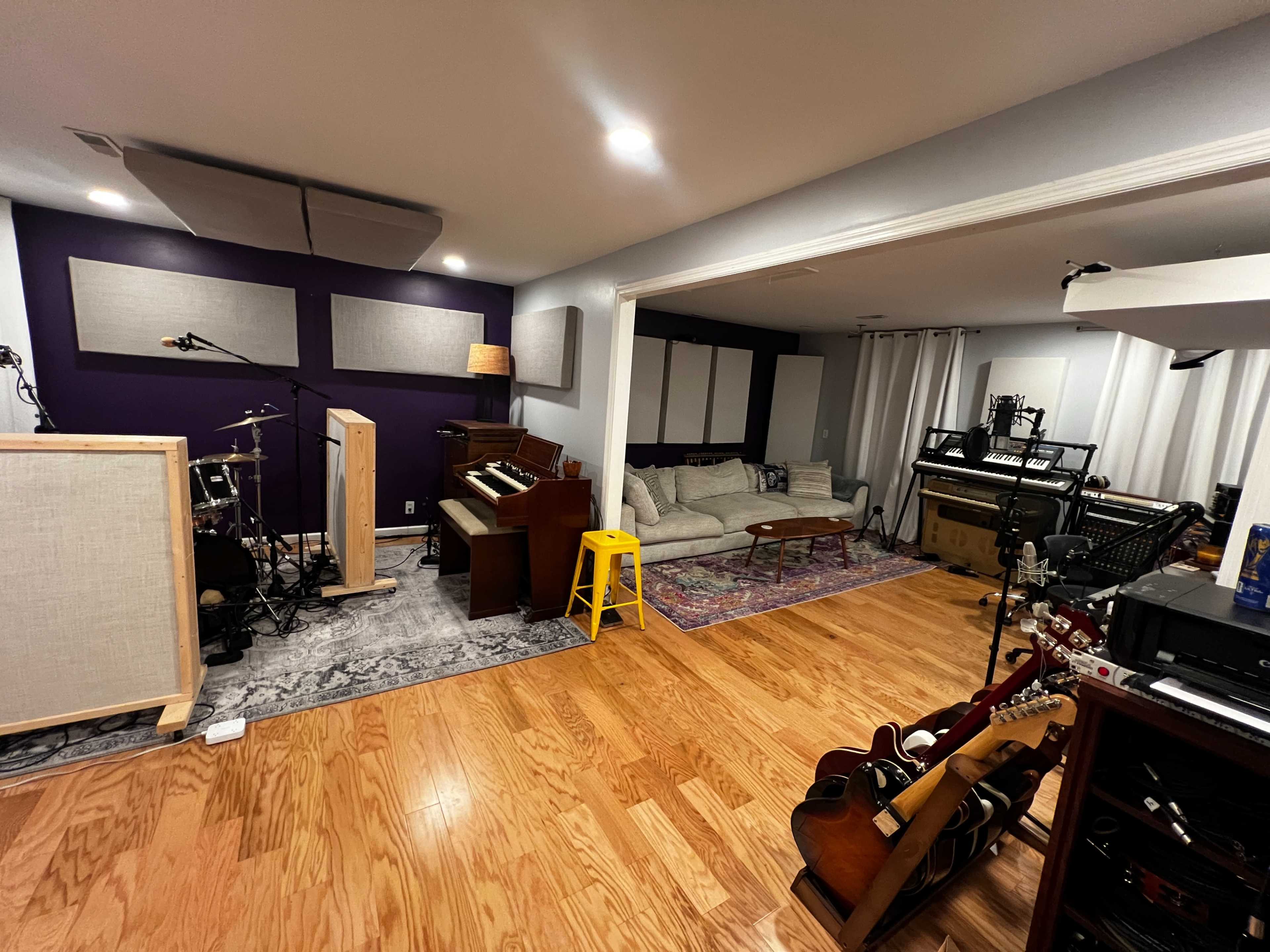 The image shows a spacious music studio featuring various instruments, a beige couch, and wooden flooring.