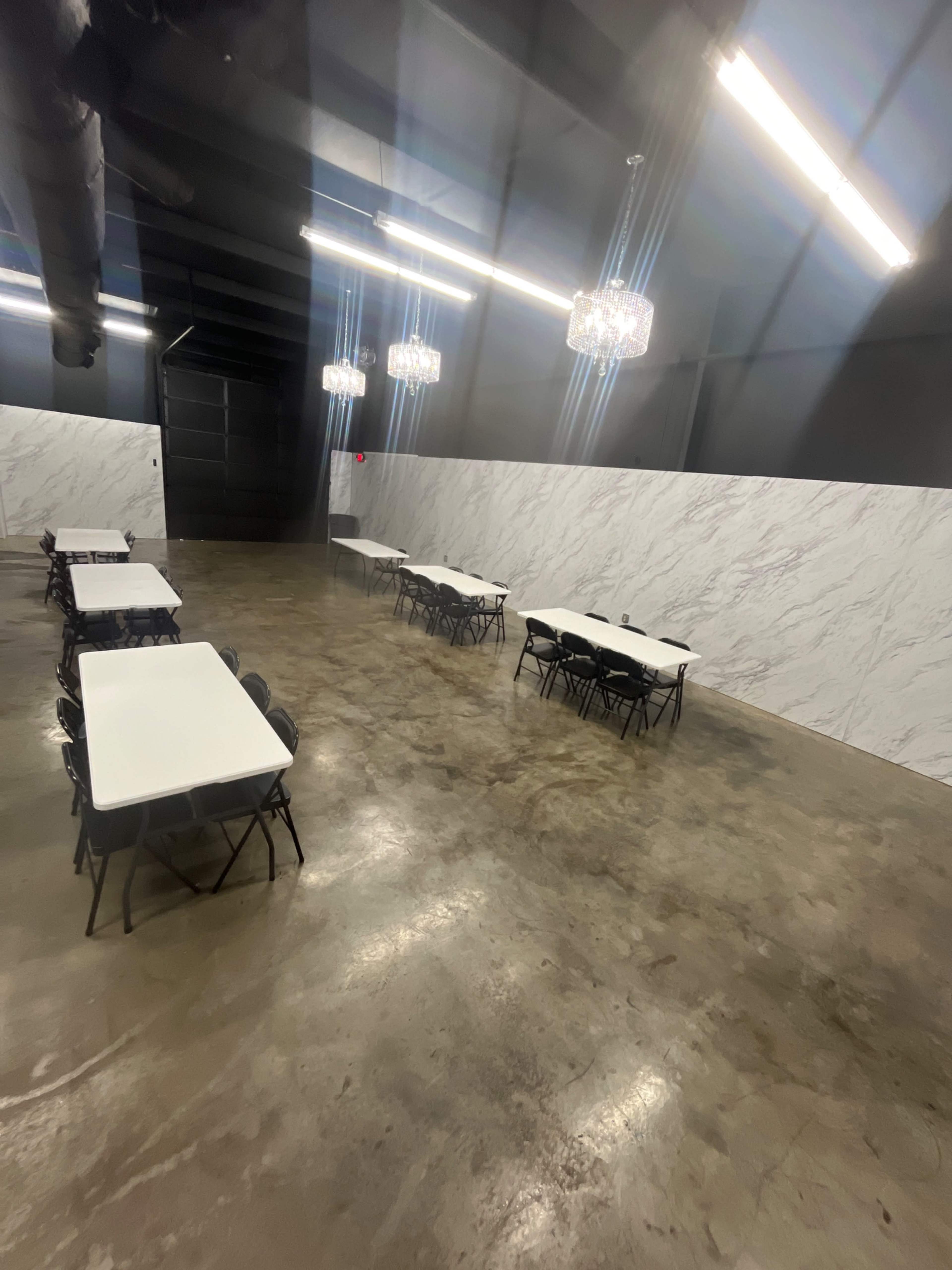 An empty room features several tables with chairs set up, surrounded by a marbled wall and illuminated by overhead lights.