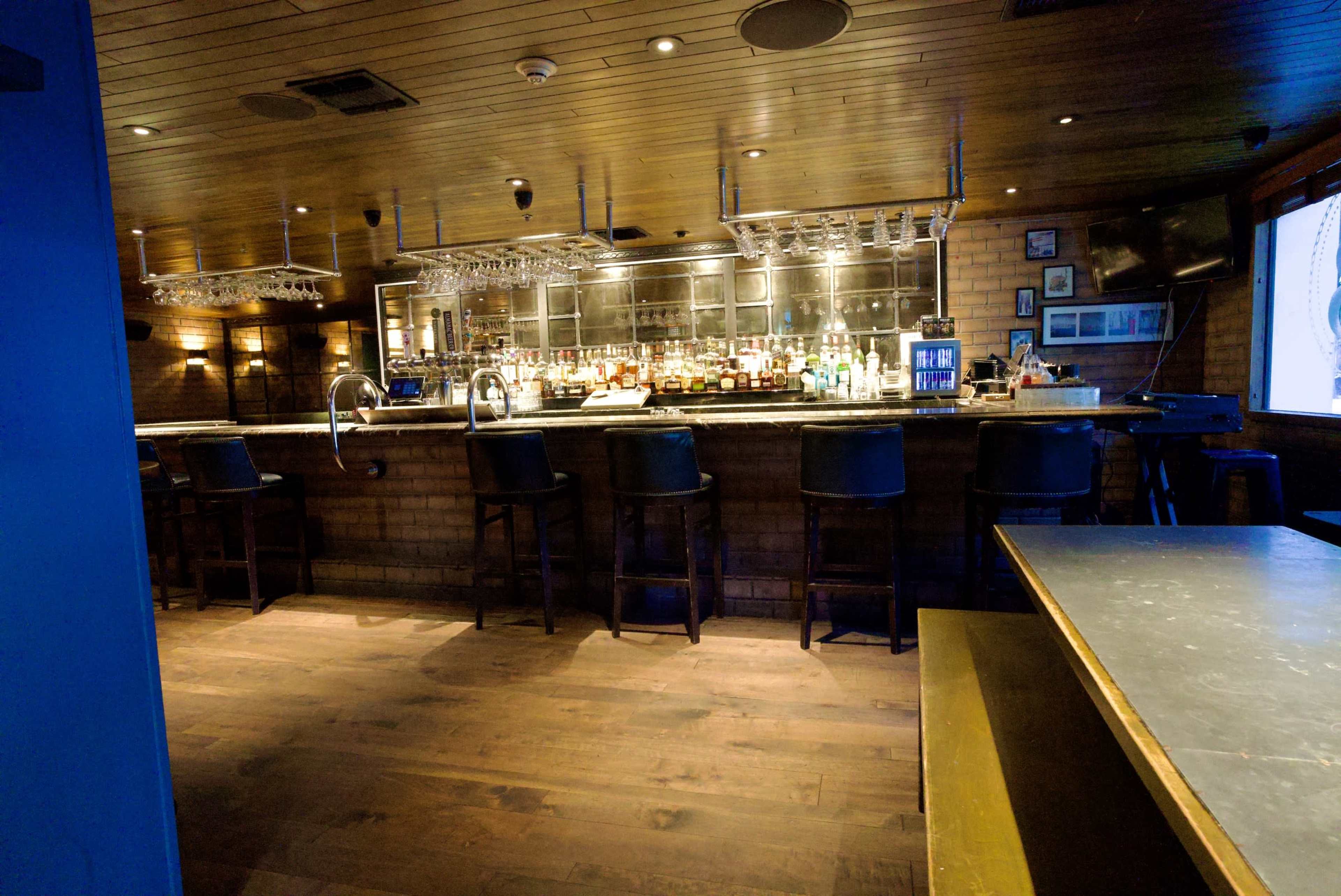 The image shows a dimly lit bar area with a wooden floor, featuring a long counter lined with bar stools and a wall of bottles behind the bar.