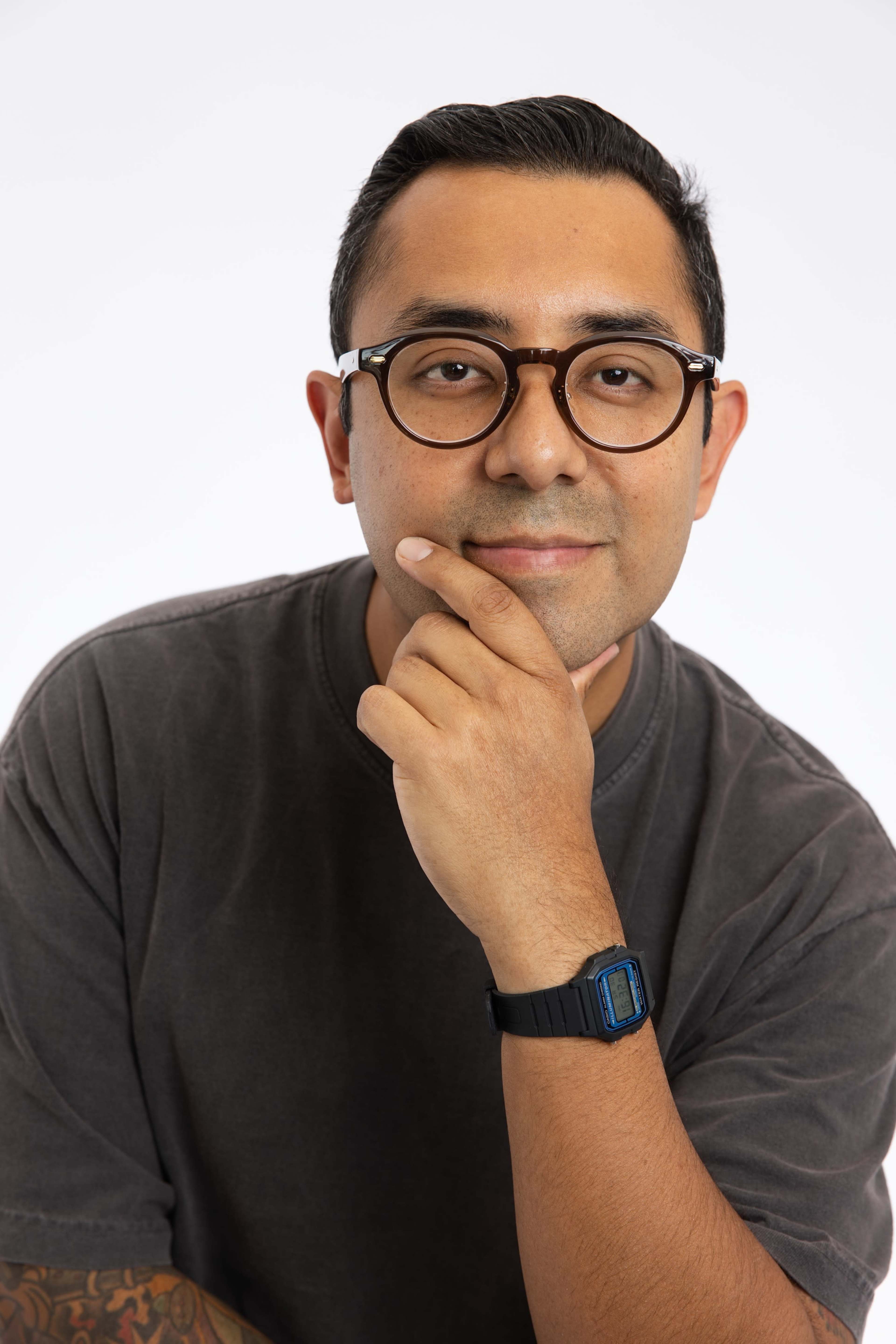 A man with glasses and a watch rests his chin on his hand against a plain white background.