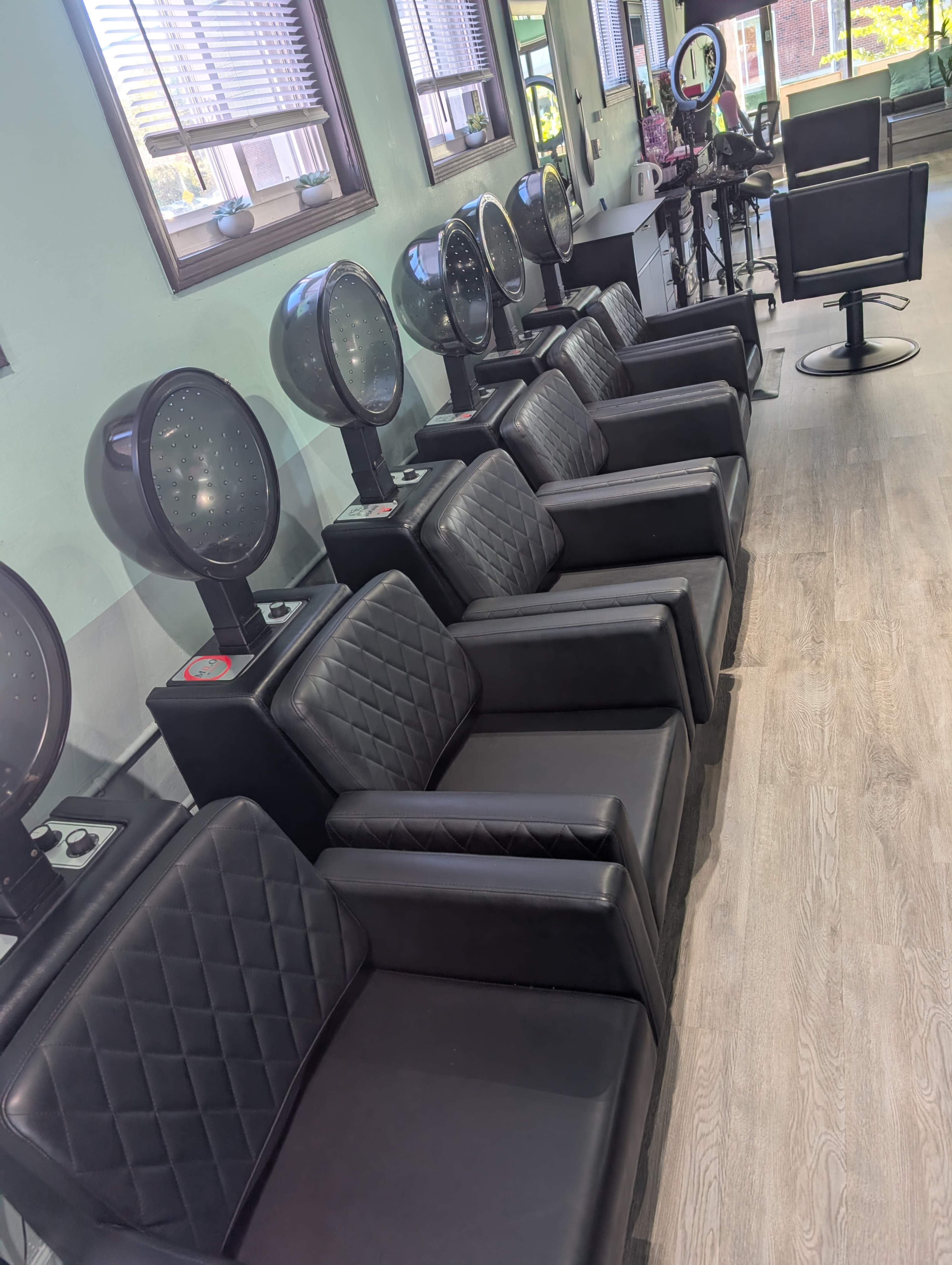 The image shows a row of black salon chairs with hooded dryers positioned against a wall in a brightly lit beauty salon.