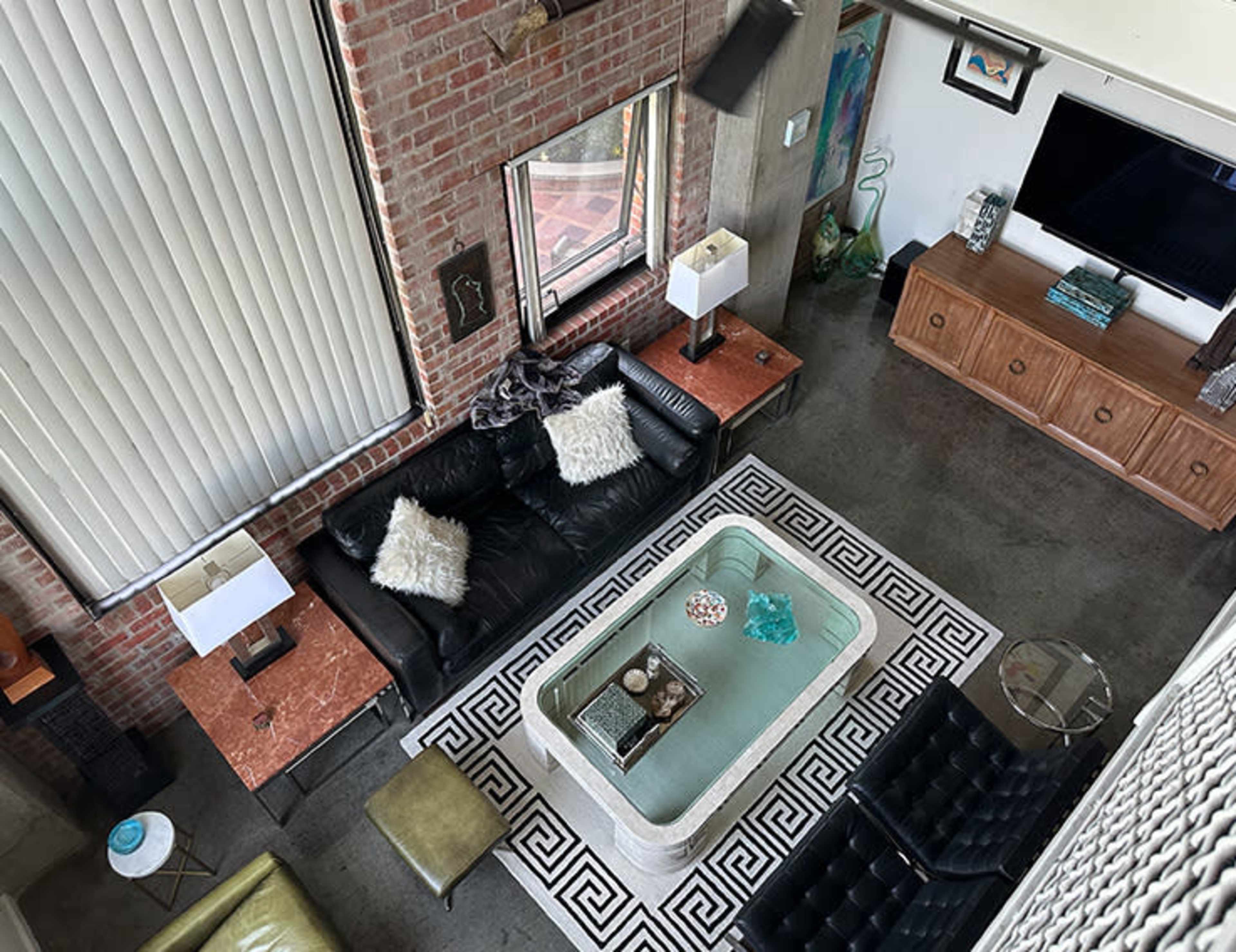 The image shows a modern living room with exposed brick walls, a black leather sofa, and a glass coffee table on a patterned rug, accompanied by side tables and a media unit.