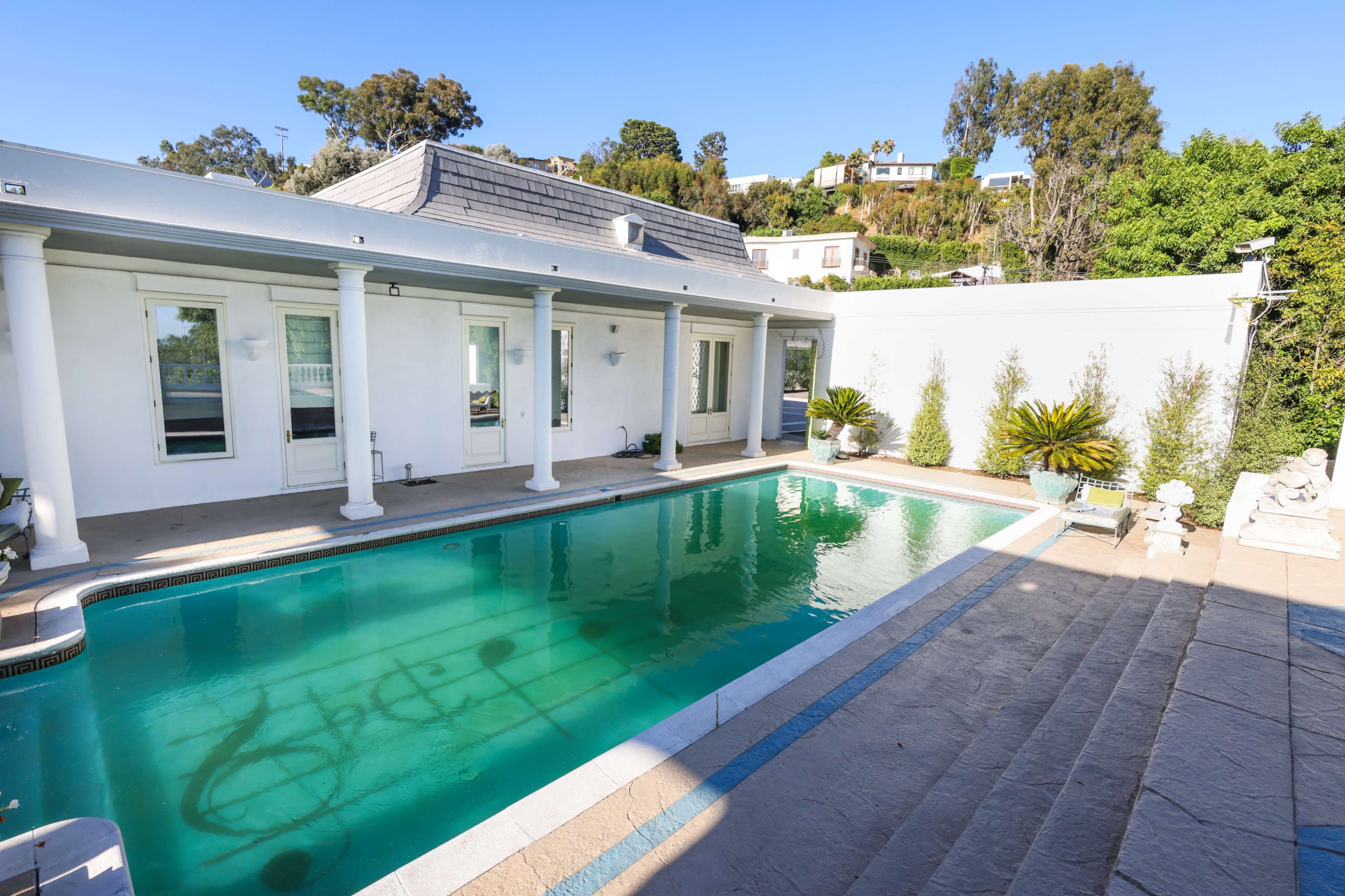 A clear blue swimming pool surrounded by a white house, stone steps, and greenery.