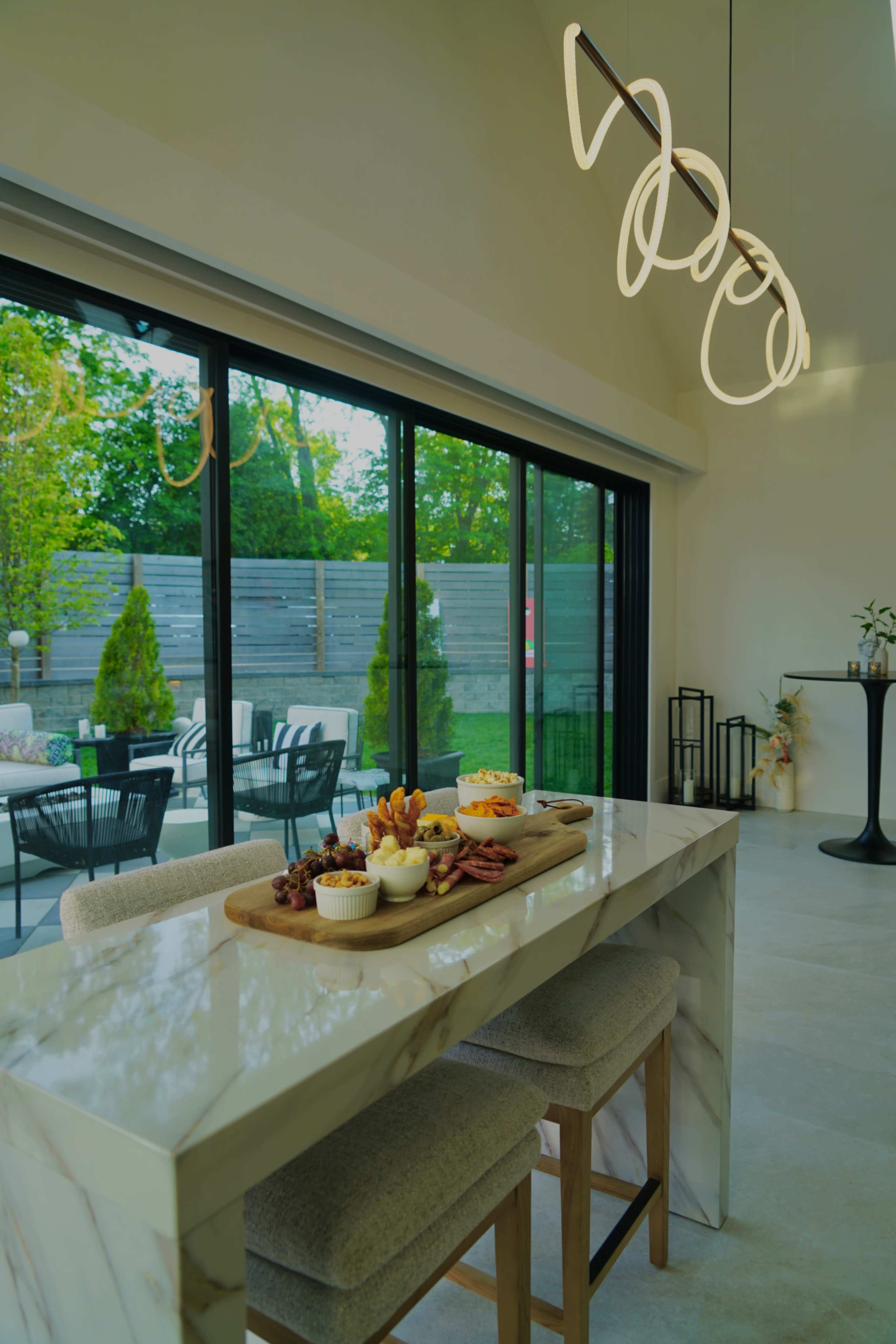 A modern kitchen features a marble countertop with a wooden serving board of snacks and a large glass door leading to an outdoor seating area.