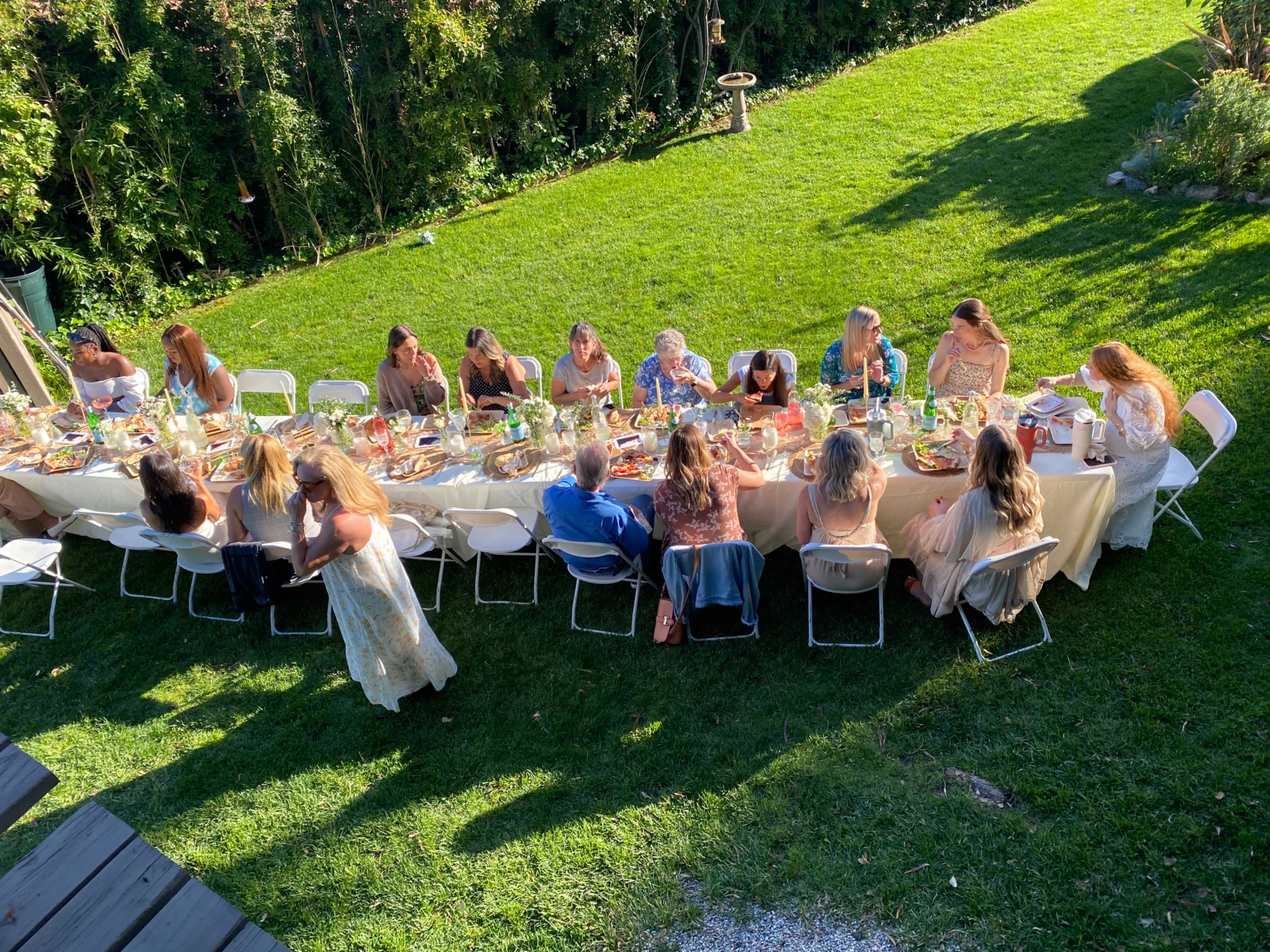 A long table is set for a gathering of people enjoying a meal outdoors on a grassy area.
