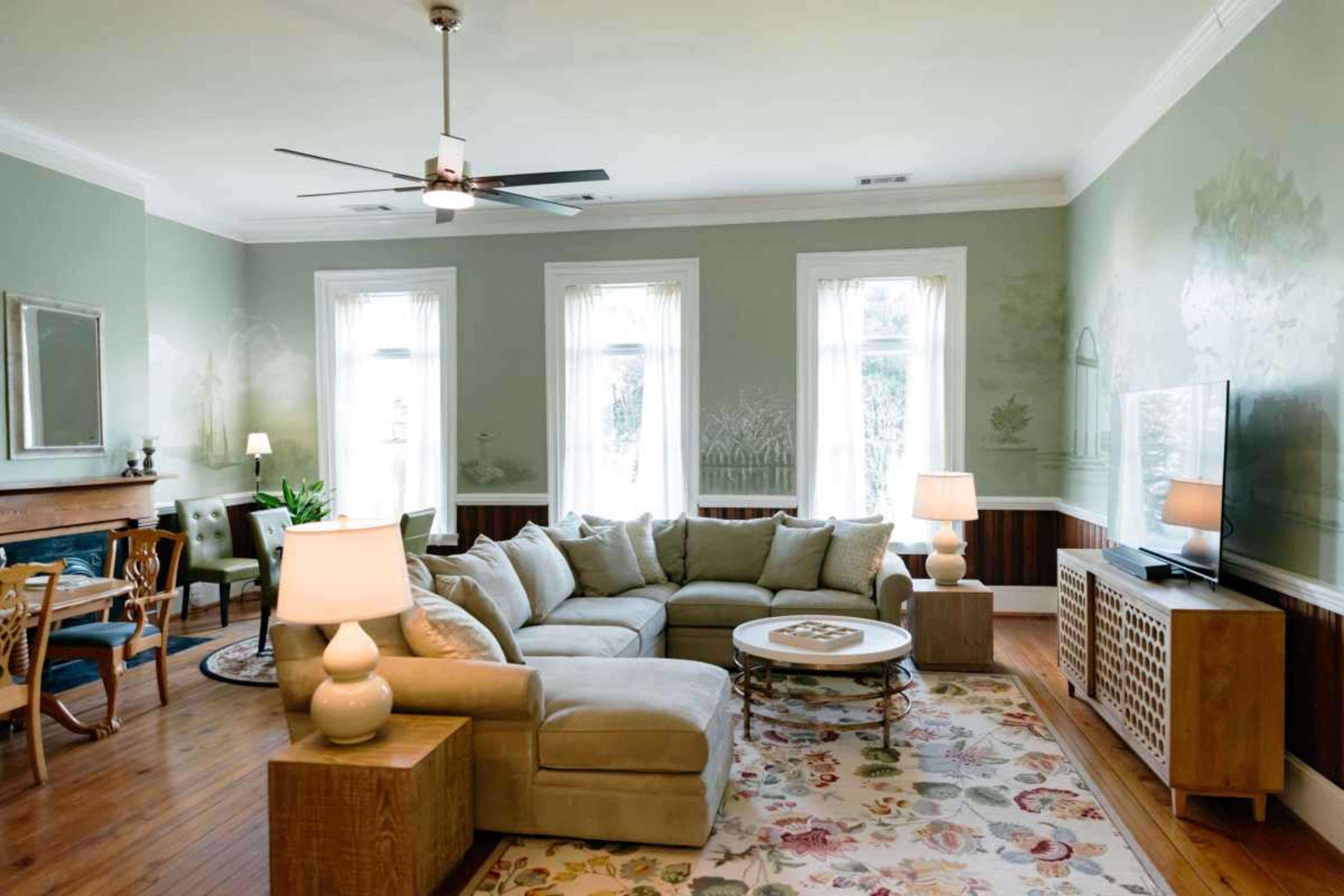 The image shows a well-lit living room with a large sectional sofa, a round coffee table, and multiple windows allowing natural light to stream in.