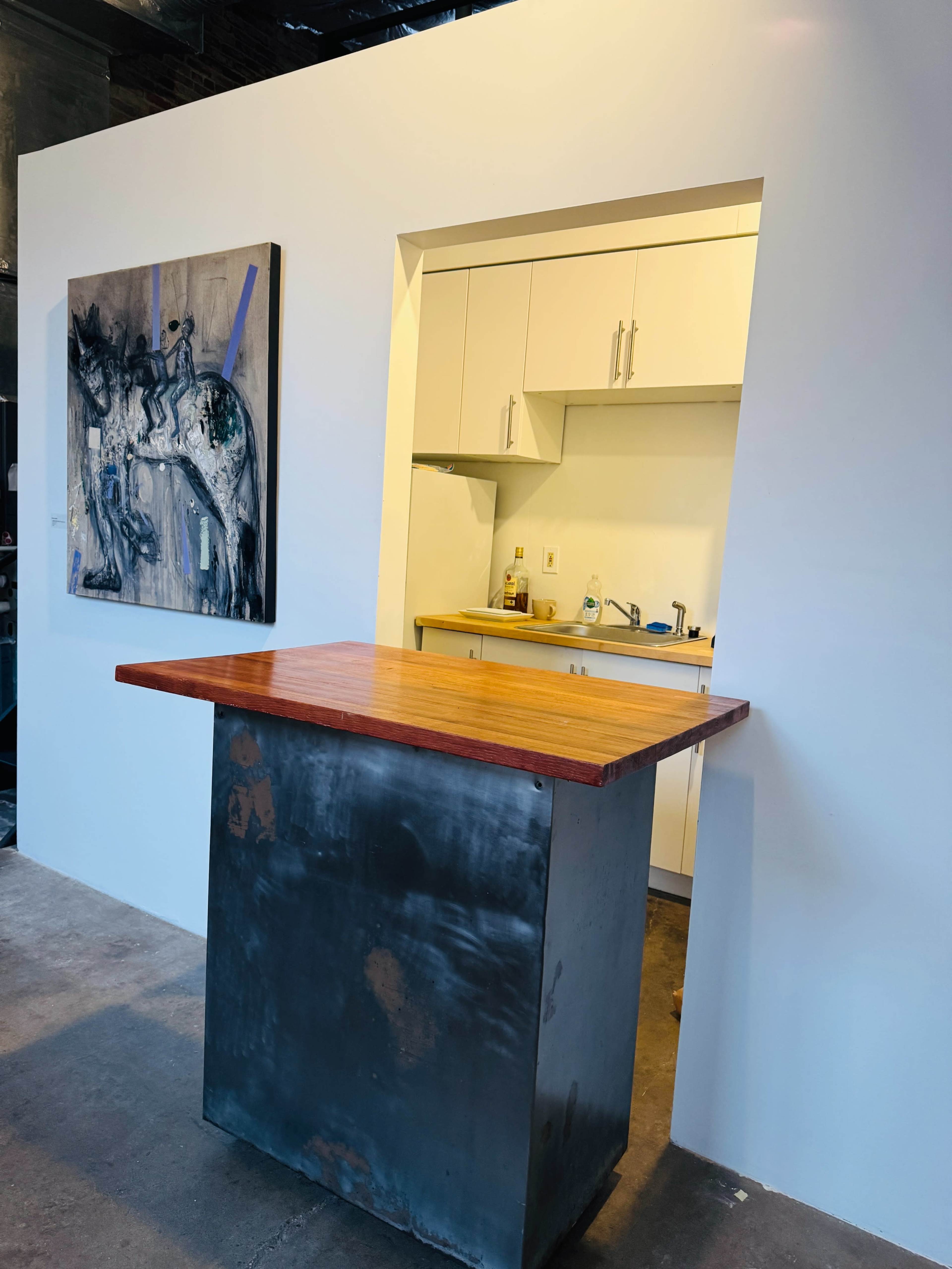 The image shows an industrial-style kitchen with a wooden countertop island and an open wall leading to a small cooking area with white cabinets.