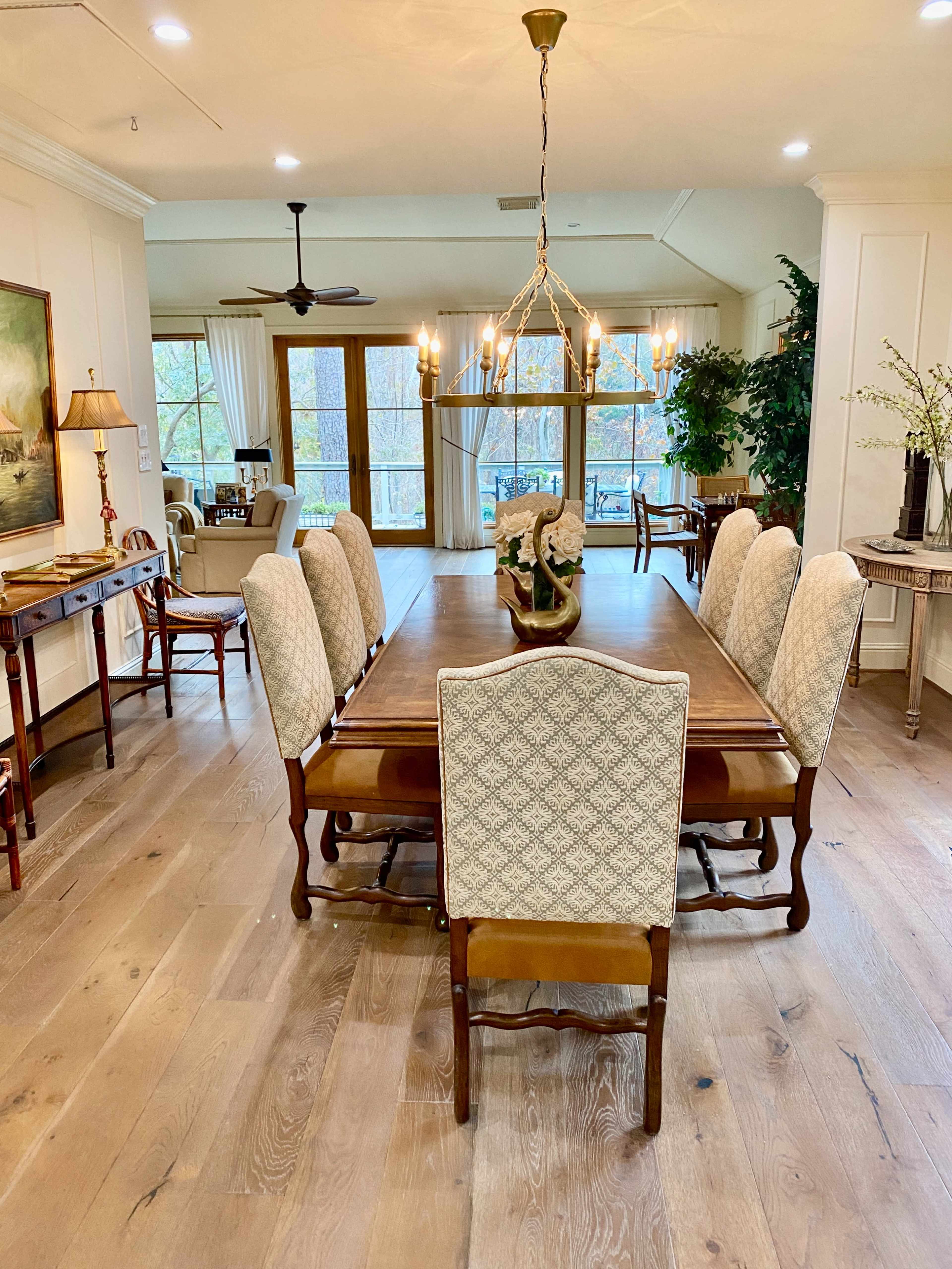 A rectangular dining table is set in a room with large windows, surrounded by eight upholstered chairs, with a chandelier hanging overhead and a view of a living area in the background.