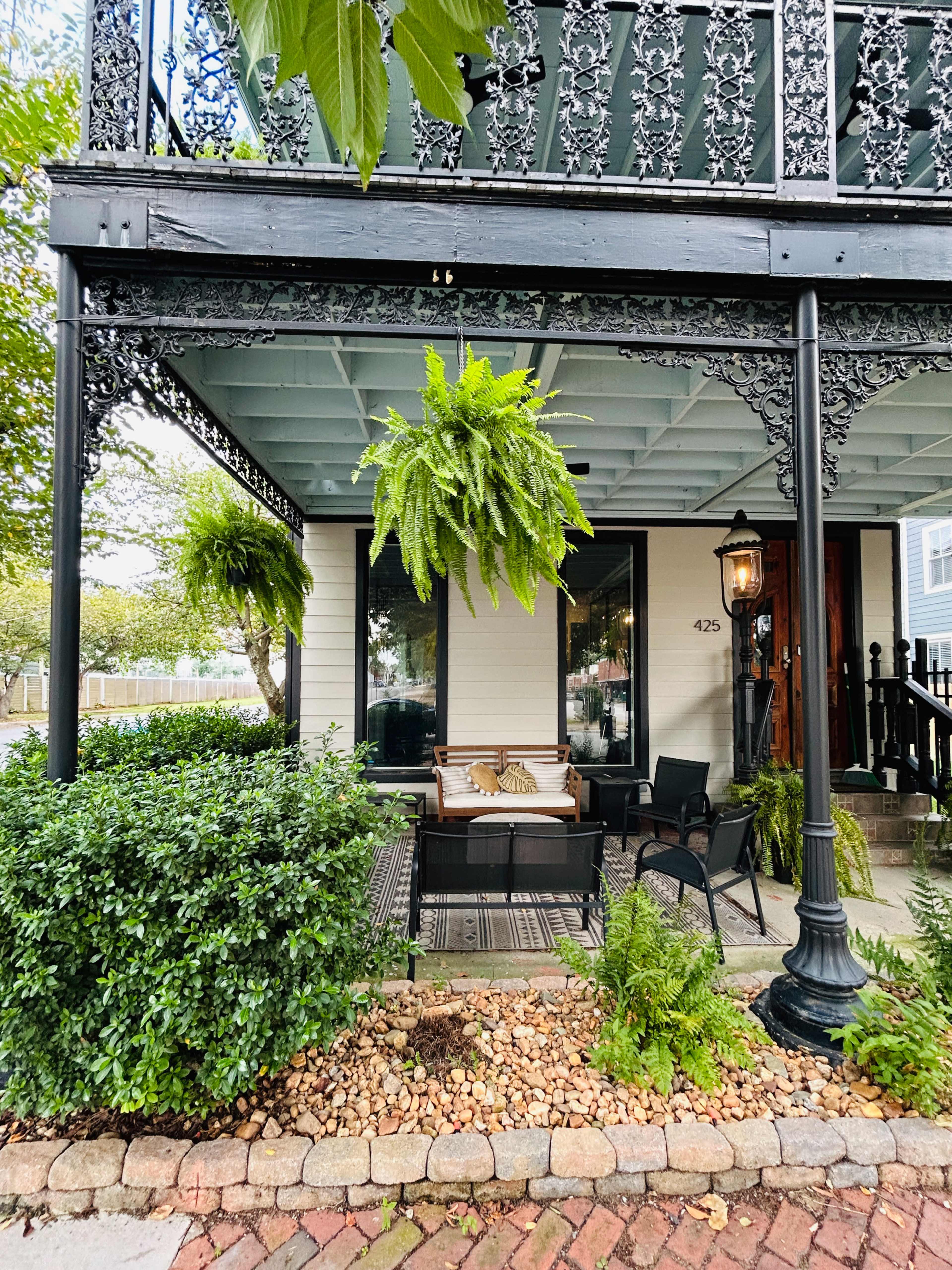 The image shows a porch with decorative ironwork, potted ferns hanging from the ceiling, a seating area with a couch and chairs, and a brick walkway lined with small stones.