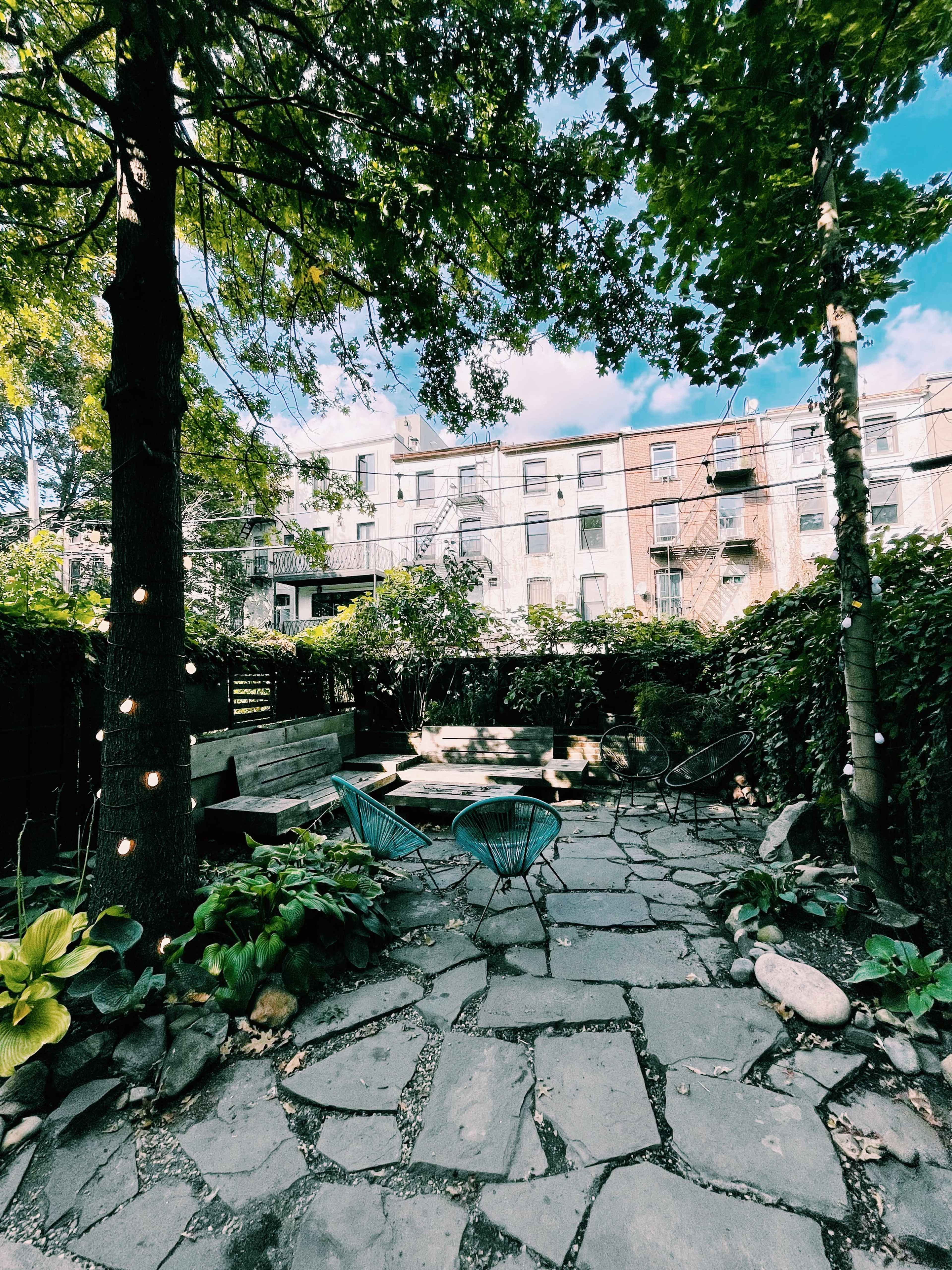 A stone-paved backyard garden with green foliage, trees, and a building with balconies in the background.