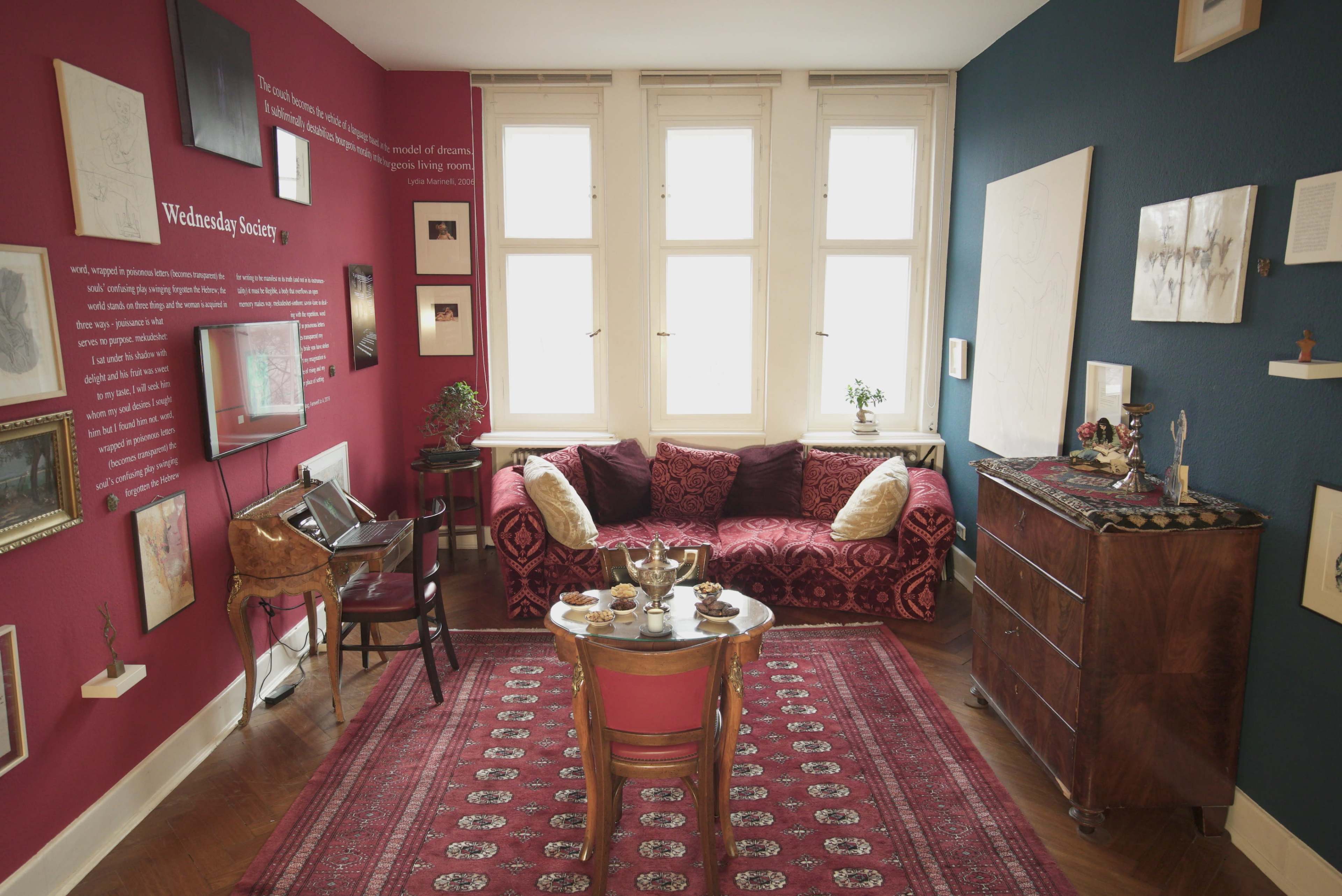 The image shows a cozy living room with a red and blue color scheme, featuring a sofa, a small table with tea elements, an area rug, and several framed artworks on the walls.