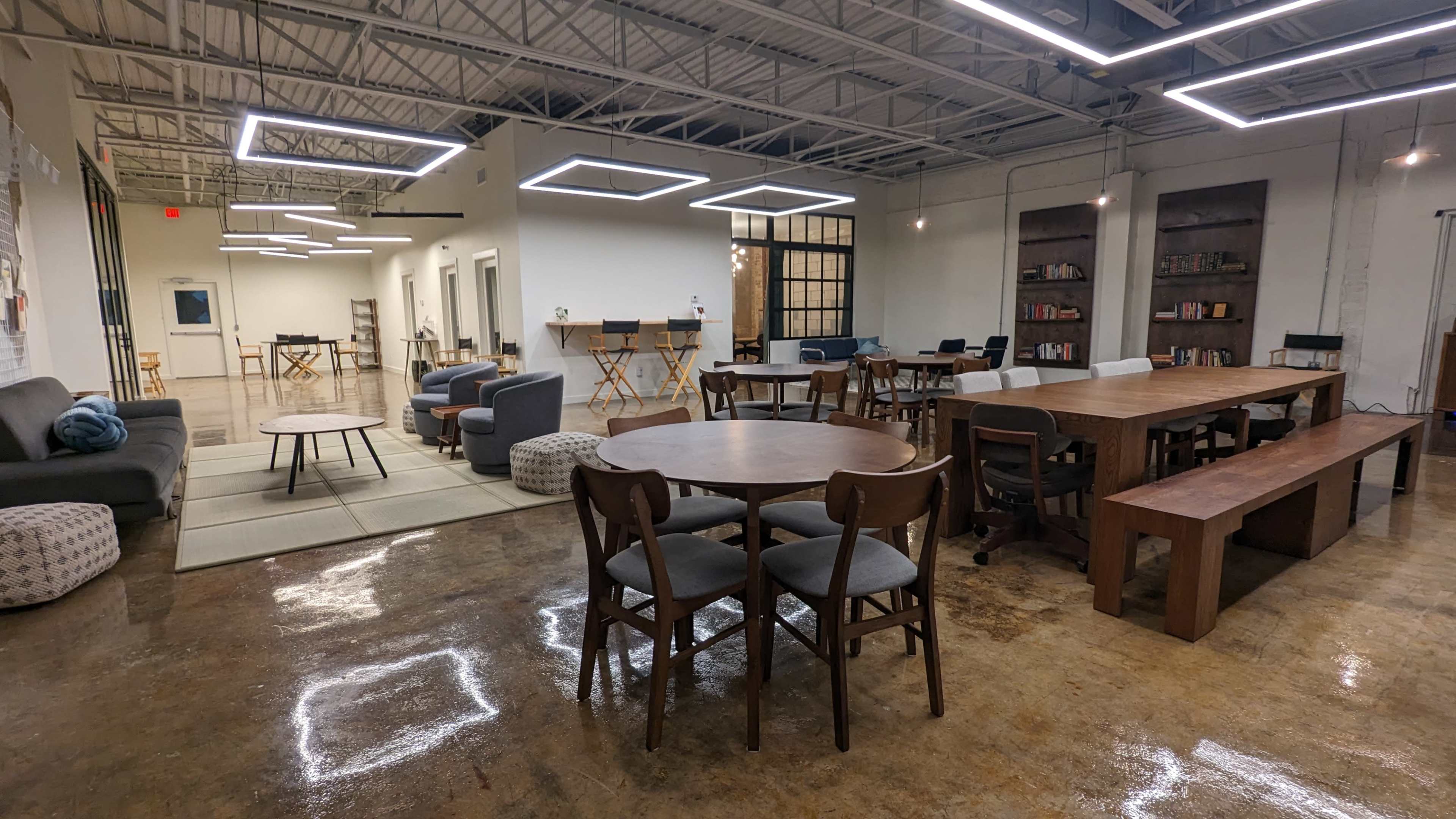 The image shows a modern, well-lit co-working space with various seating arrangements, including tables, chairs, and couches, along with shelves of books.