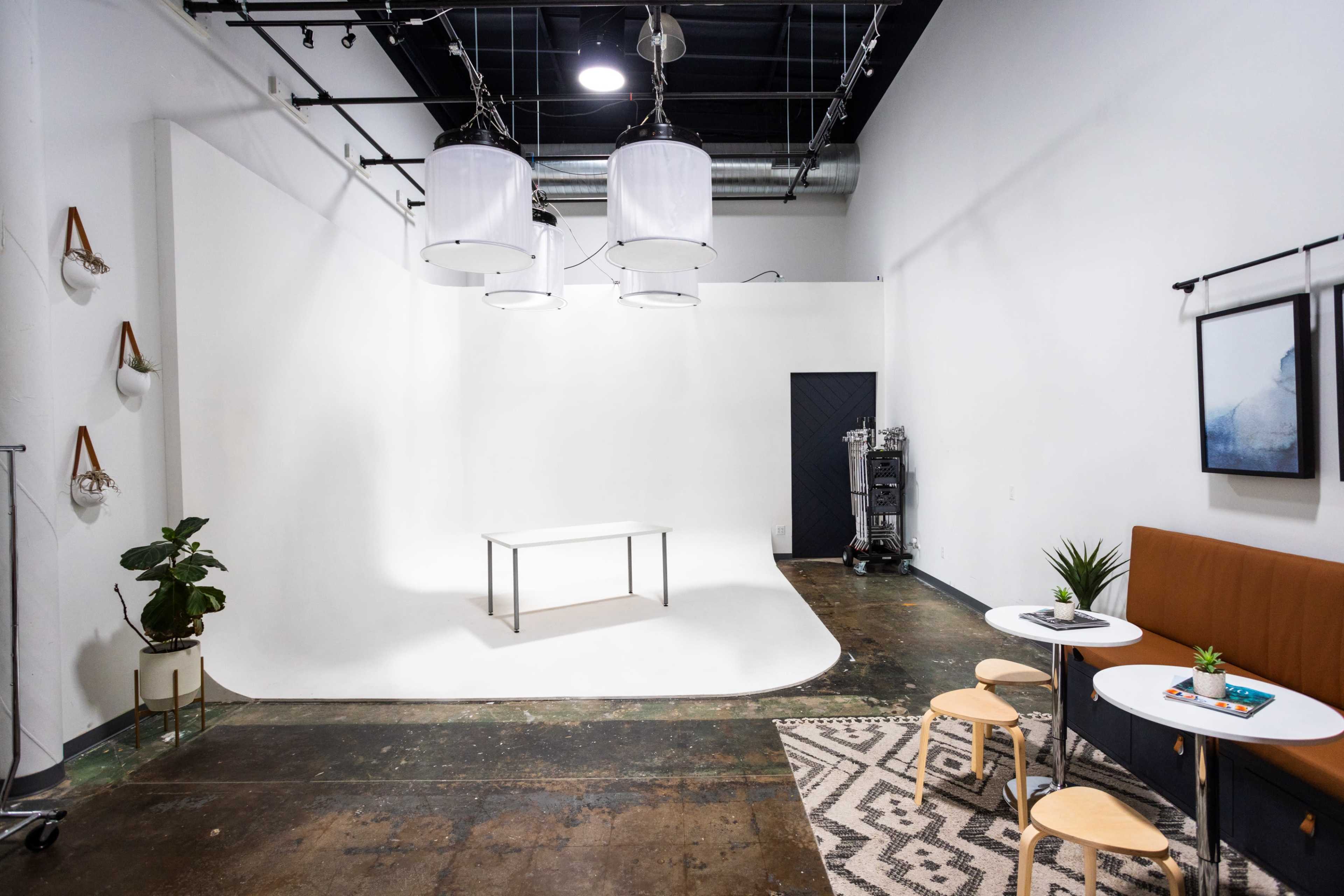 The image shows a minimalist photography studio with three hanging lights, a central table, and a seating area featuring a small table and chairs.