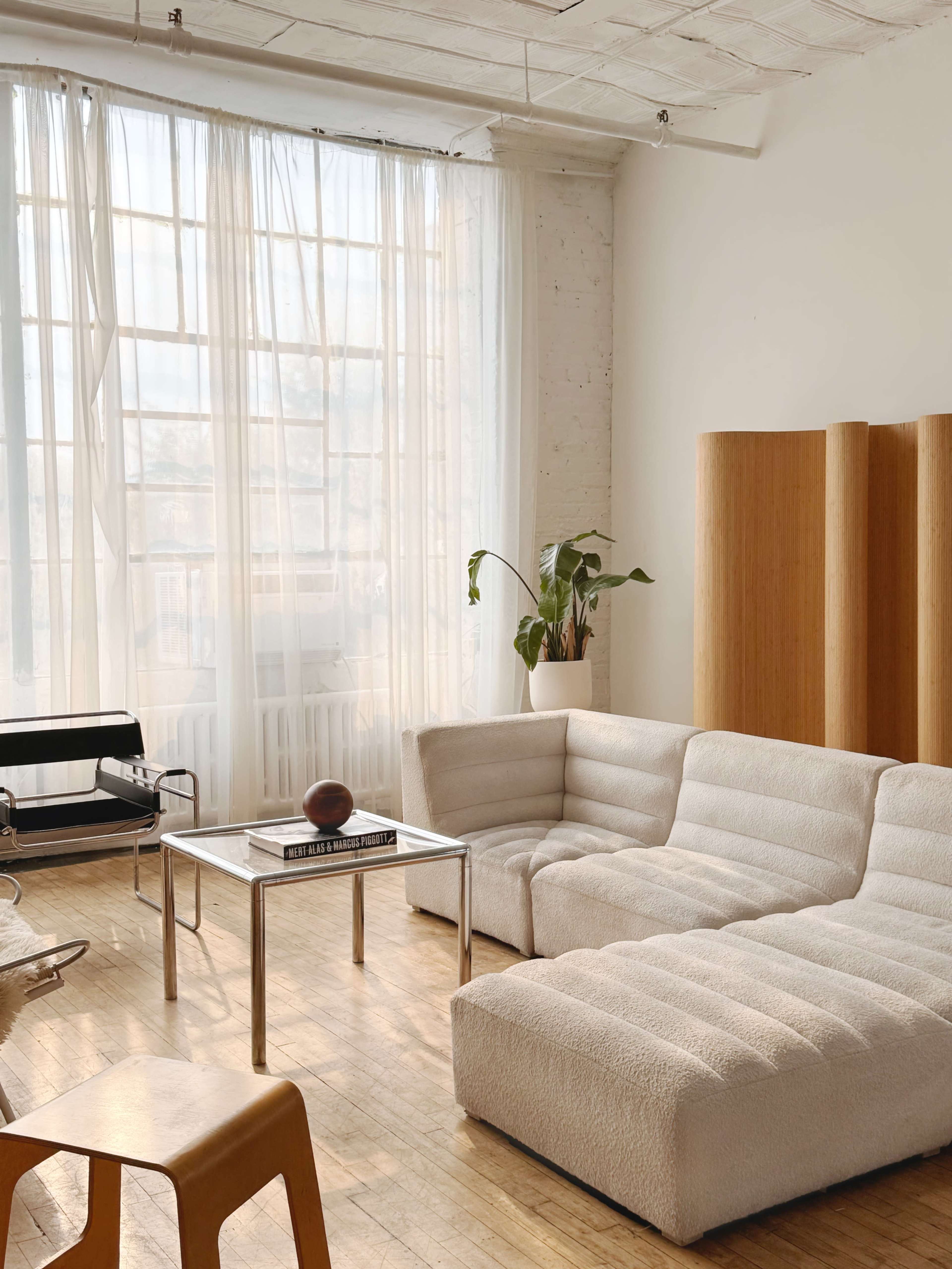 The image shows a bright, minimalistic living room featuring a large, white upholstered sectional sofa, a glass coffee table, and wooden room dividers against a backdrop of large windows with sheer curtains.
