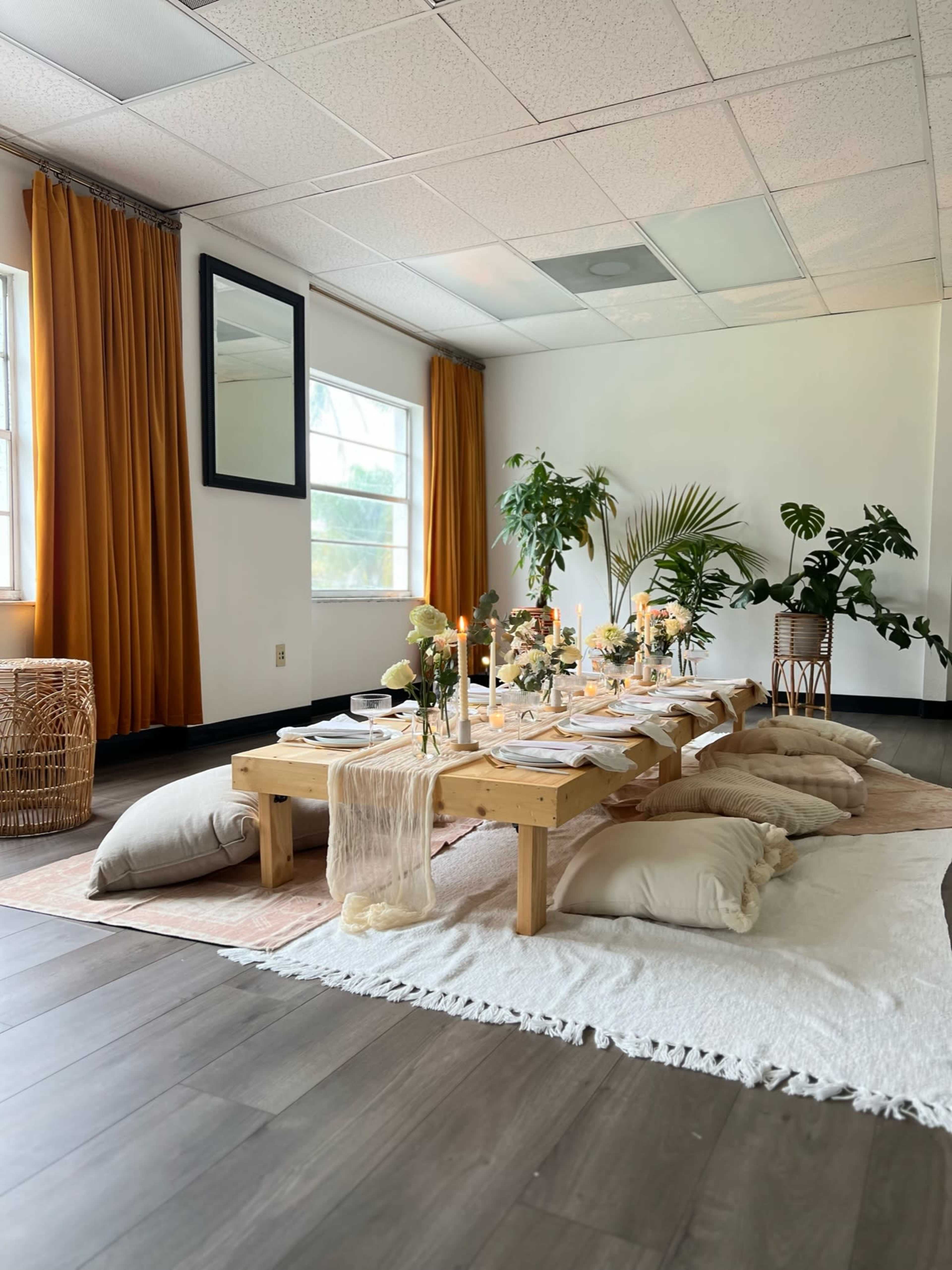 A dining area features a low wooden table set with dishes and floral centerpieces, surrounded by cushions on a decorative rug.