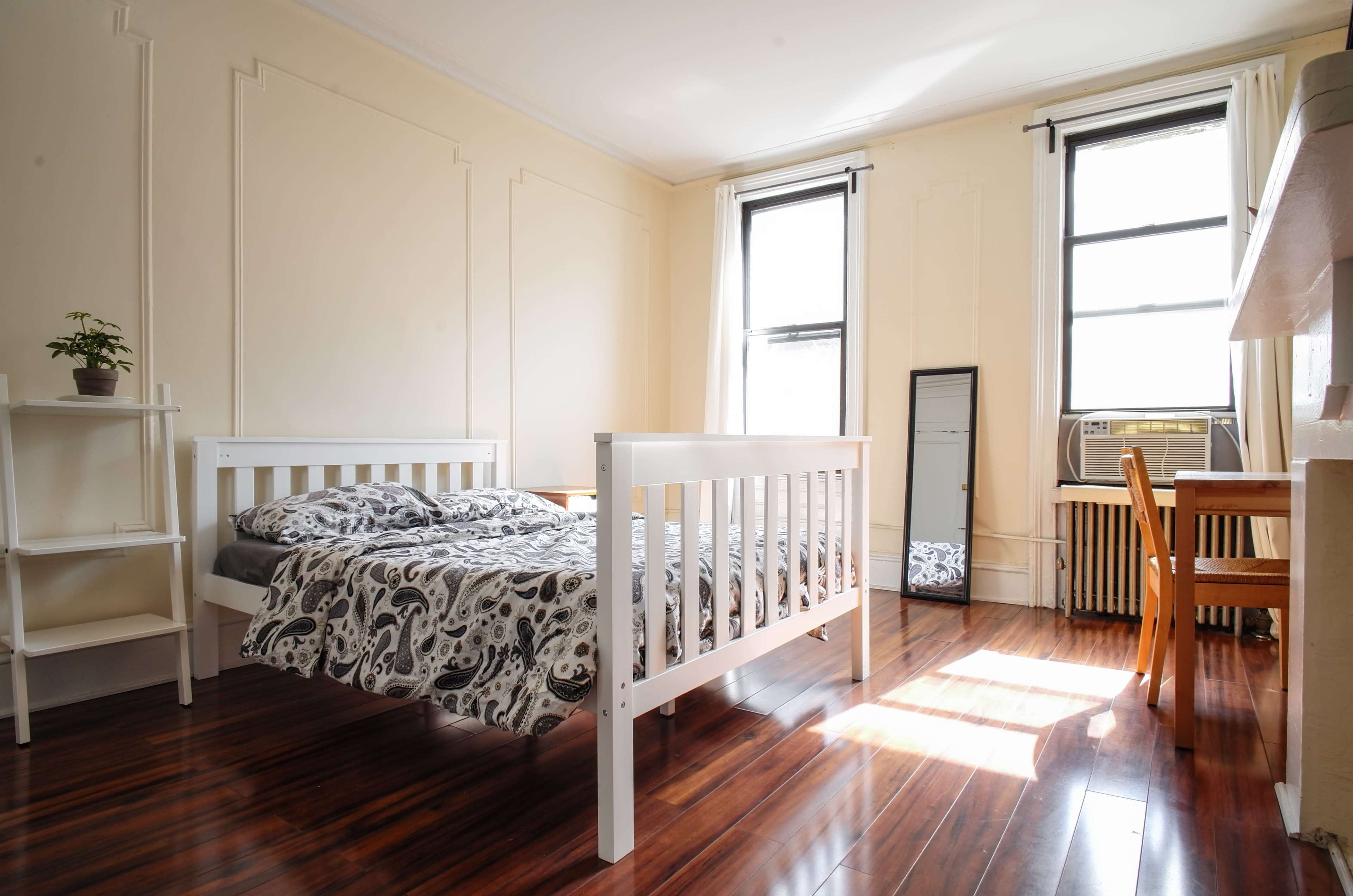 A brightly lit bedroom featuring a white bed with patterned bedding, a small potted plant on a shelf, a mirror, and a wooden table with a chair.
