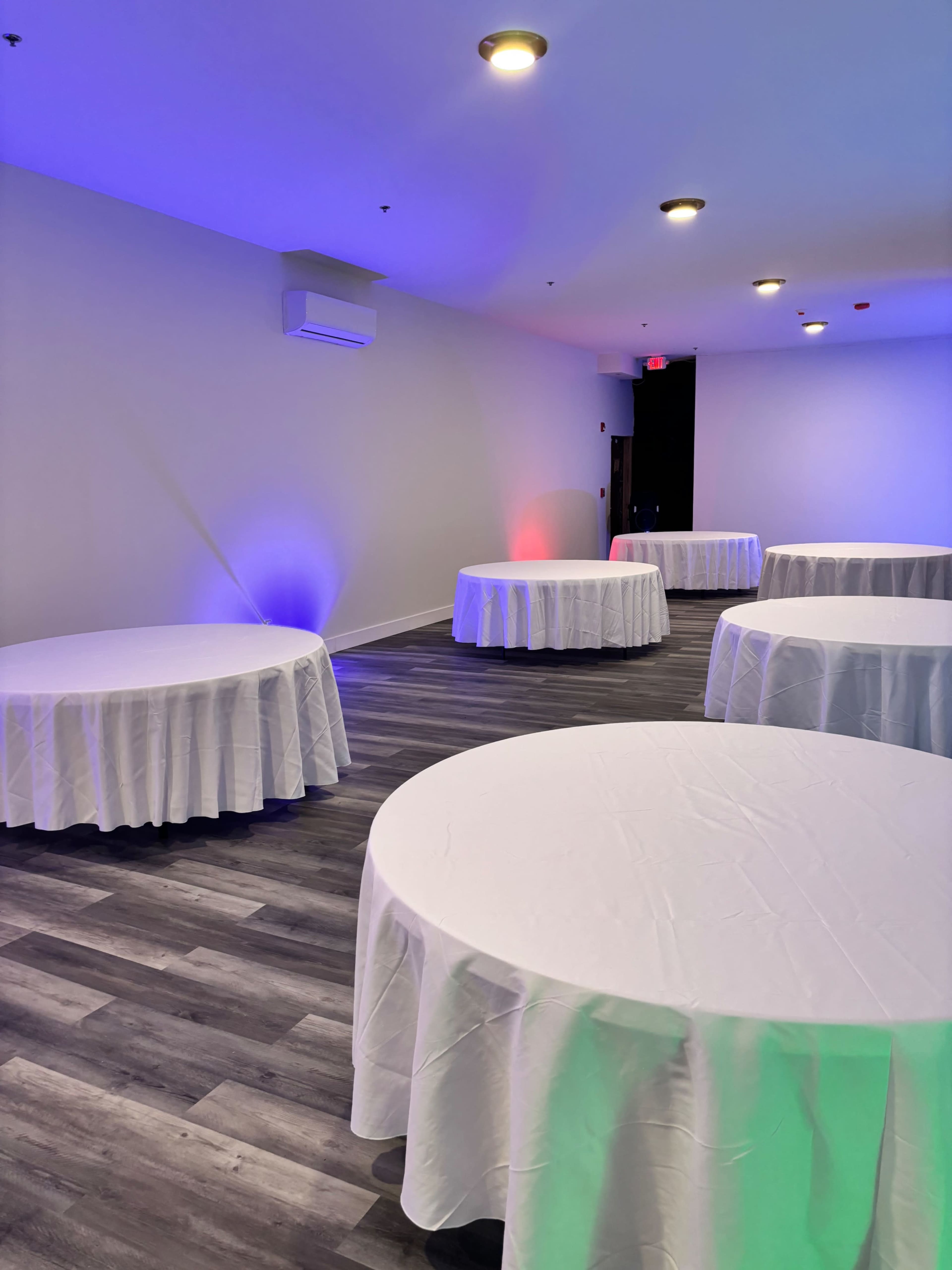 A spacious room with multiple round tables covered in white tablecloths, illuminated by colored lights along the walls.
