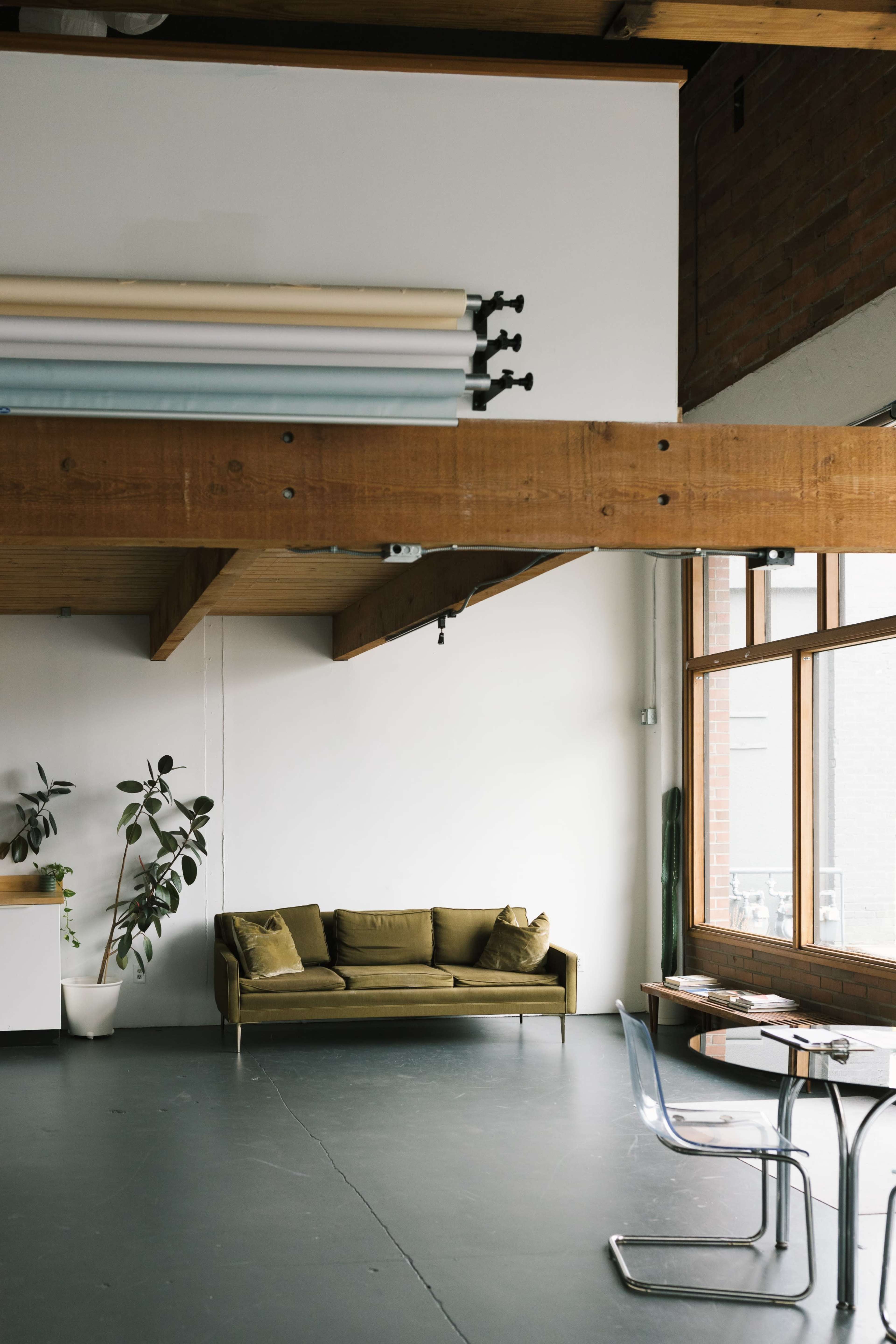 A modern living space features a green sofa, large windows, exposed wooden beams, and a potted plant.