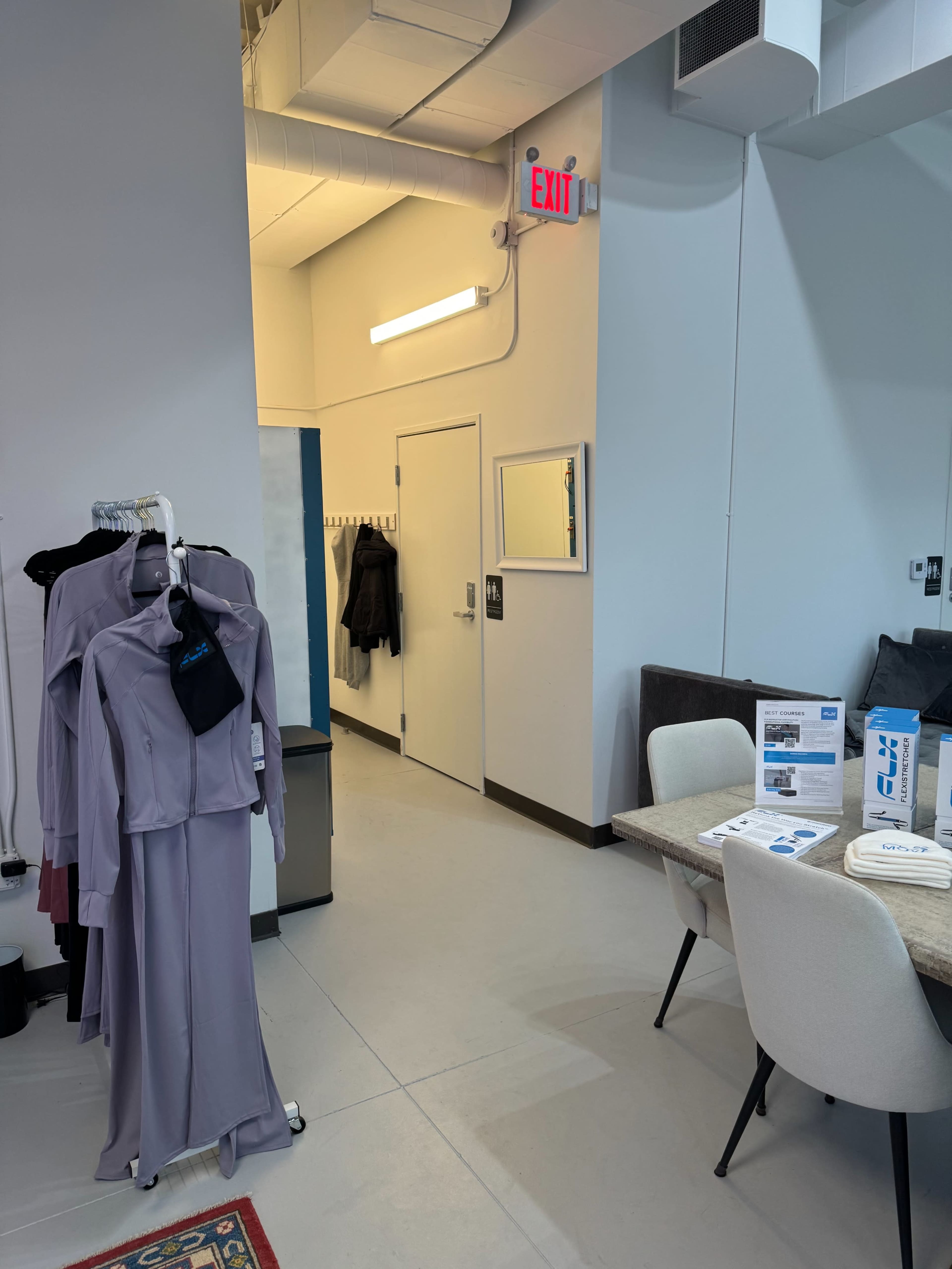 The image shows a modern interior space featuring a clothing rack with garments, a table and chairs, and a door leading to another area.