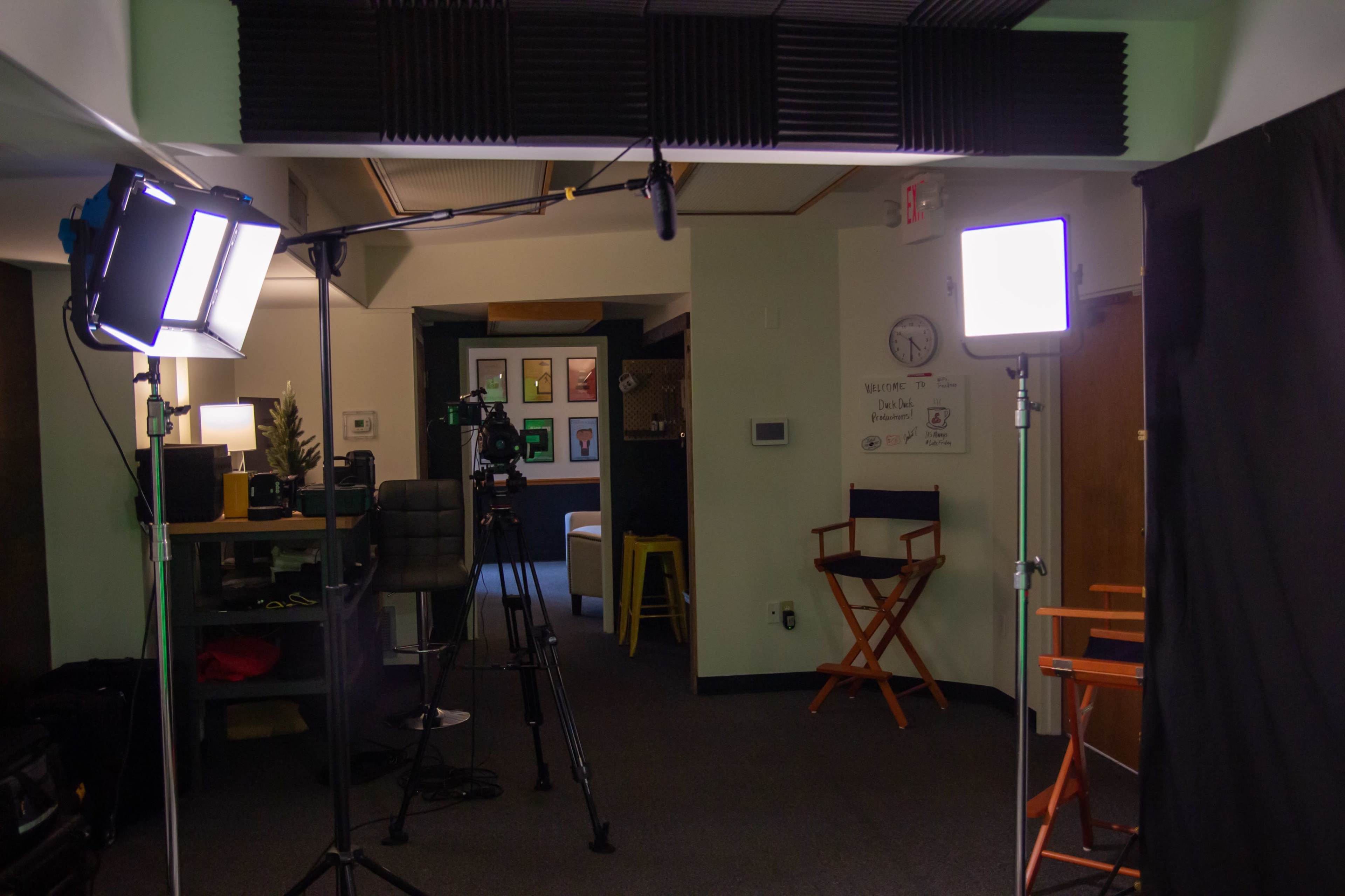 A film studio setup with two lighting stands, a camera on a tripod, and chairs positioned in front of a black backdrop.