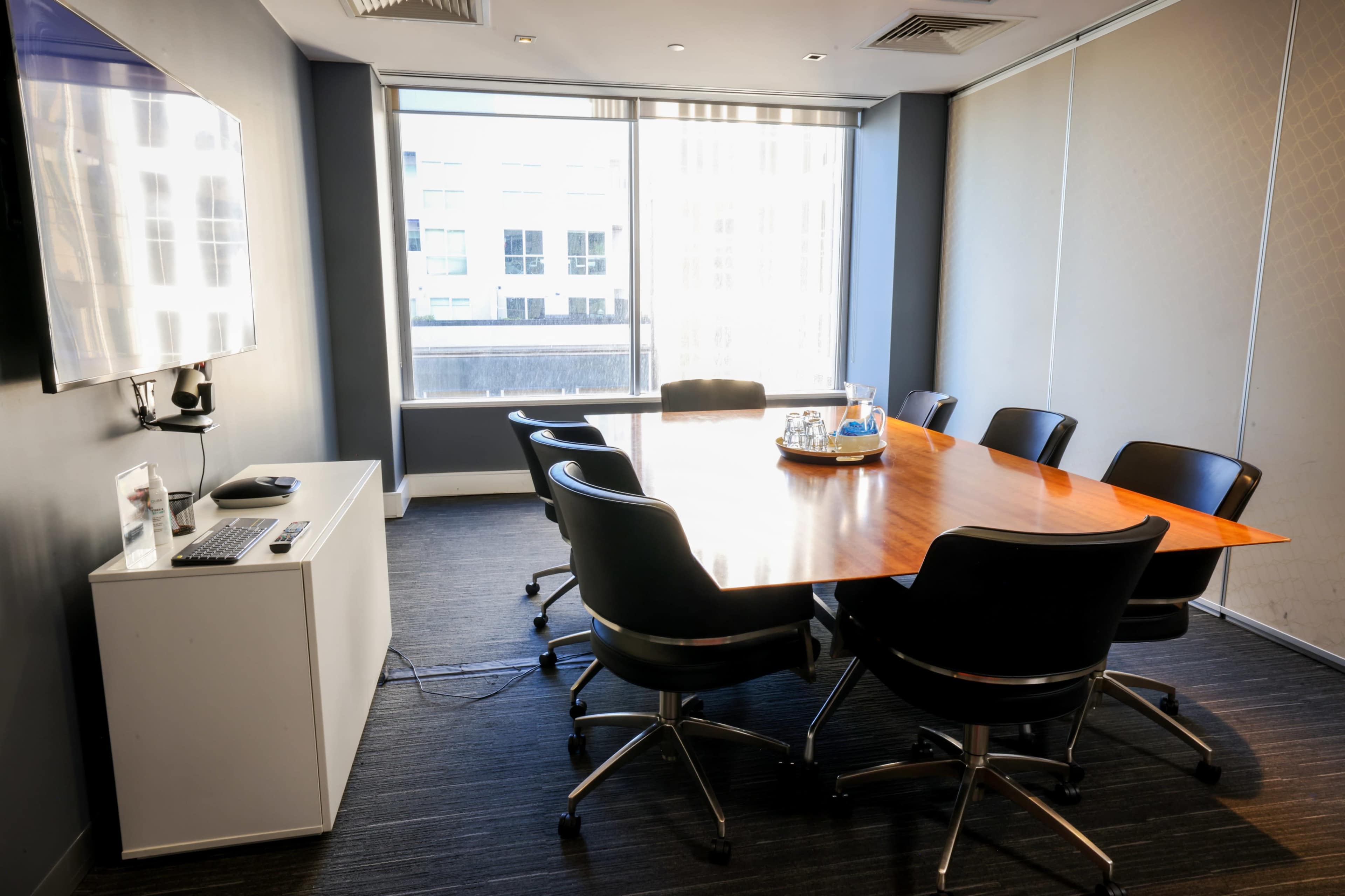 A conference room features a large wooden table surrounded by black office chairs, with a window showing a cityscape in the background.