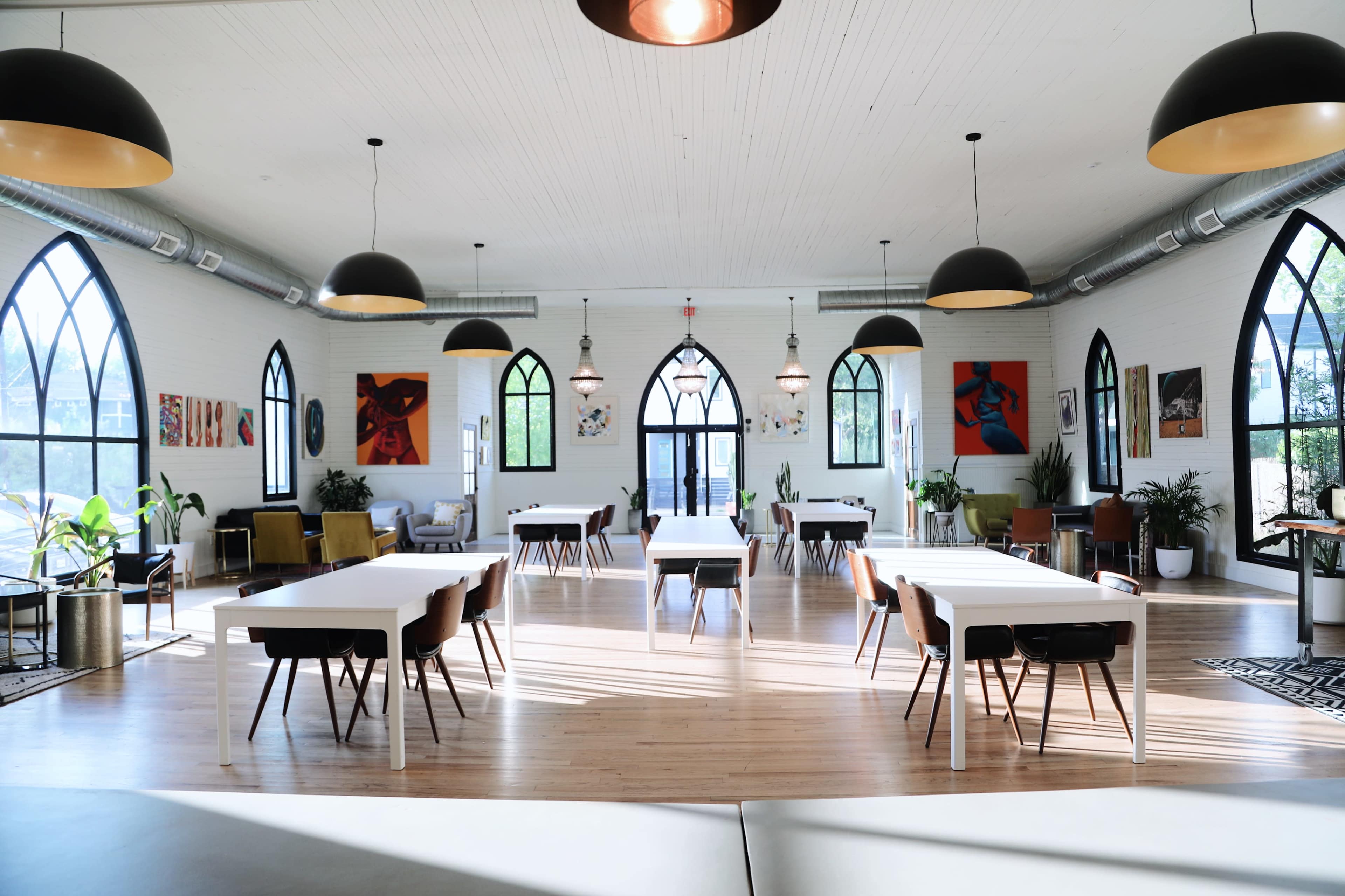 A spacious, well-lit interior of a room with high ceilings, featuring tables and chairs, large arched windows, and various artworks on the walls.