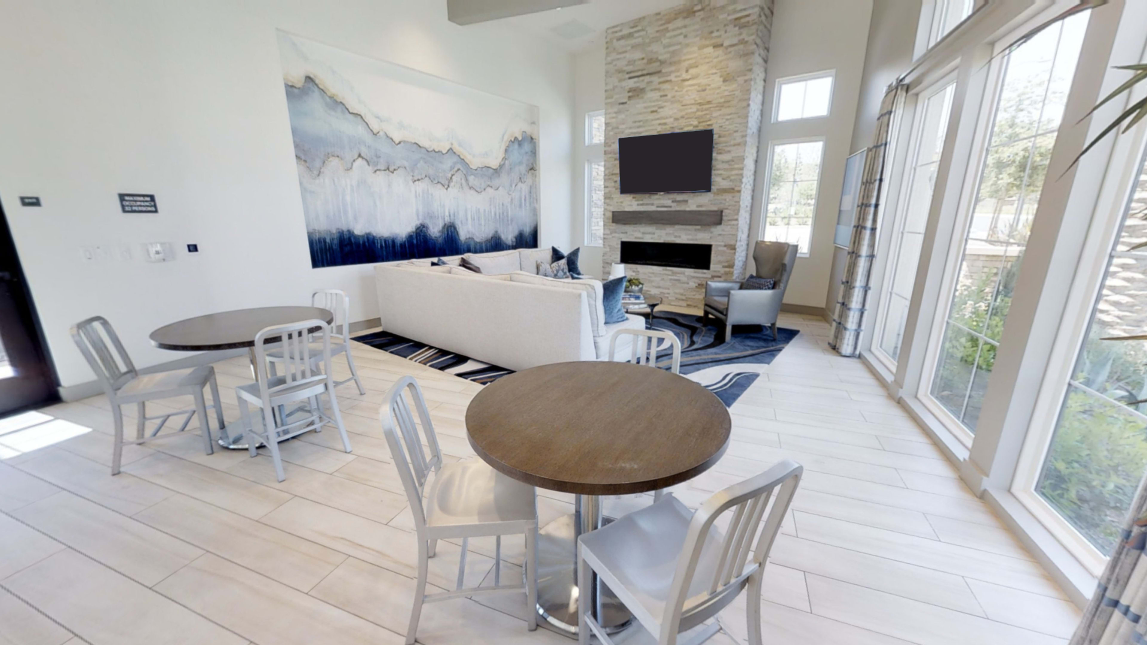 A modern lounge area with a large abstract painting, a light-colored sofa, and two tables with chairs arranged near floor-to-ceiling windows.