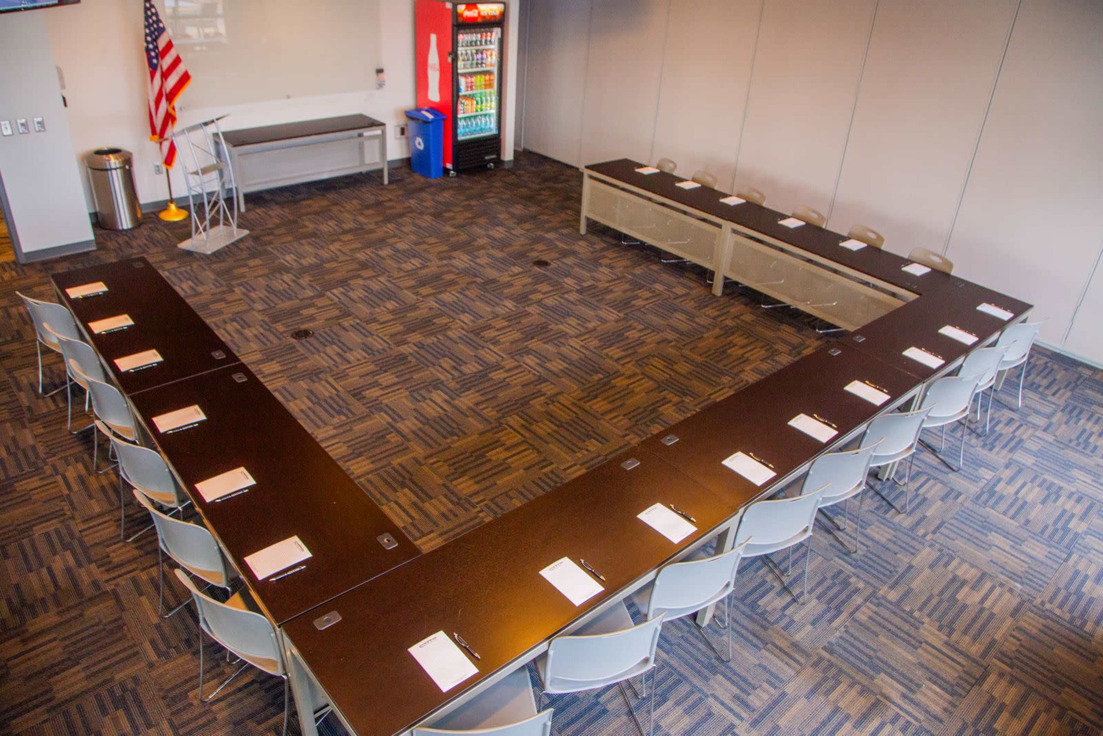 A large conference room features a U-shaped arrangement of dark tables surrounded by gray chairs, with an American flag and a vending machine in the background.