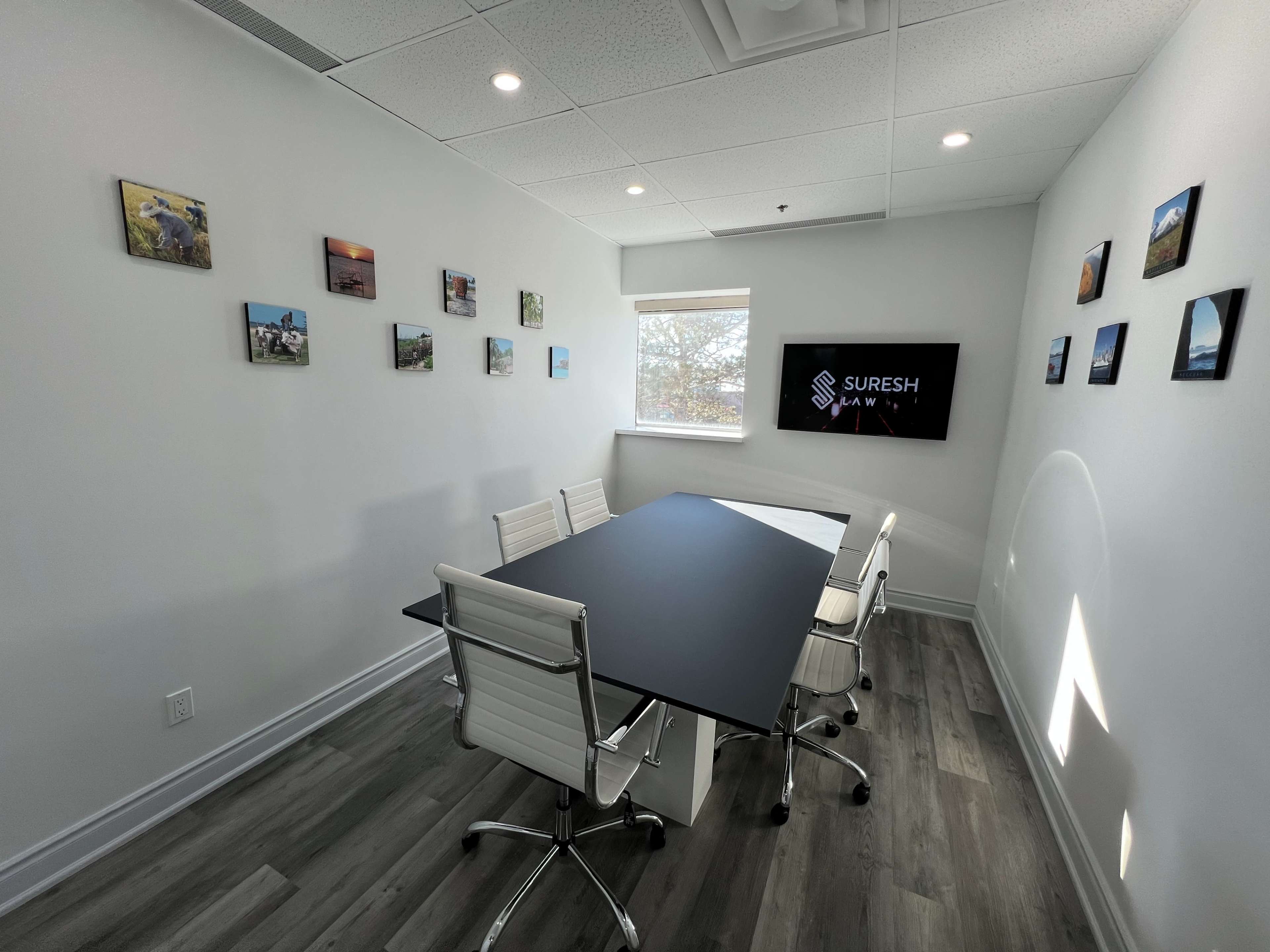 A modern conference room with a large black table, white office chairs, and framed photos hanging on the walls, along with a screen displaying the name "Suresh."