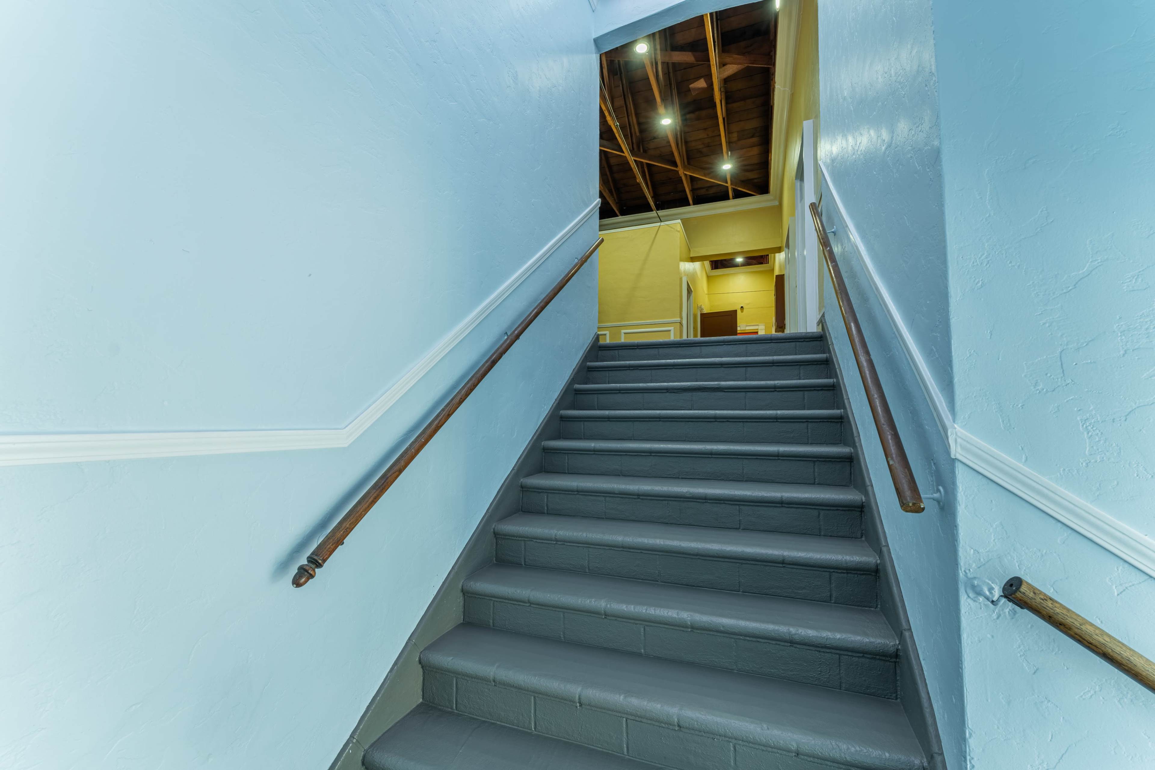A set of gray stairs leads upward, with a light blue wall and wooden handrails, opening into a well-lit area at the top.