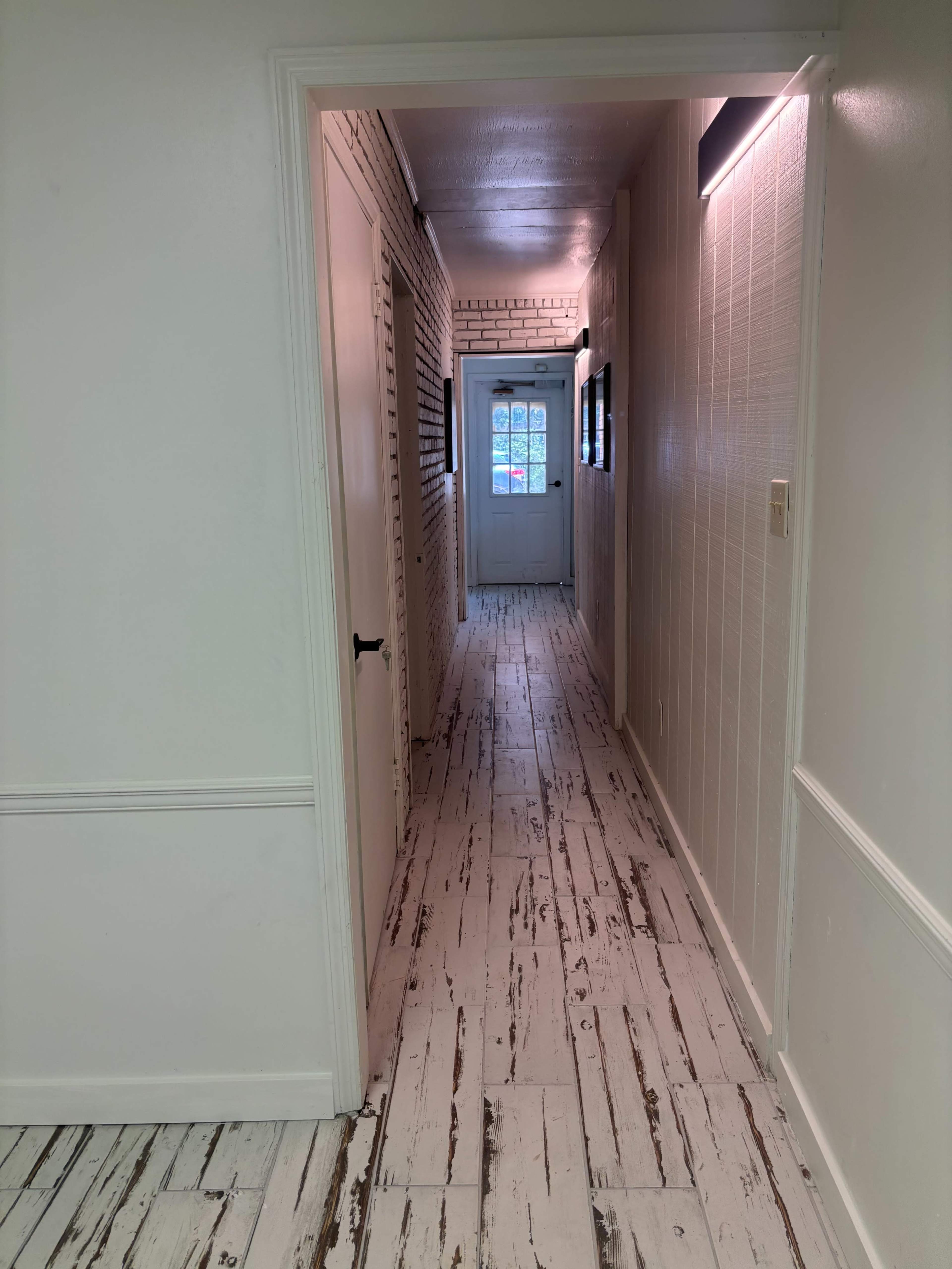 The image shows a long, narrow hallway with white plank flooring, leading to a door at the end.