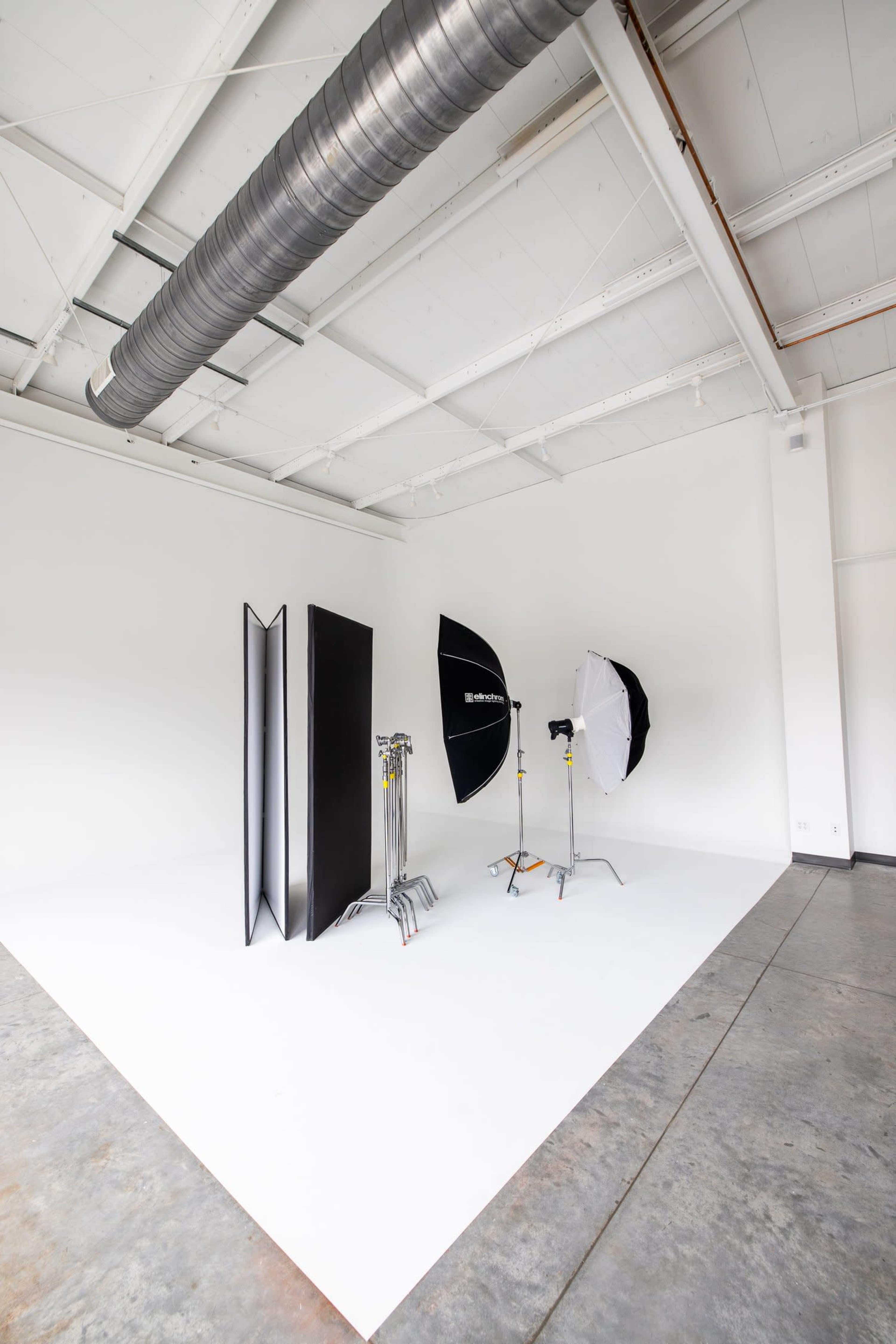 The image shows a minimalist photography studio with various lighting equipment and a backdrop setup against a white wall.