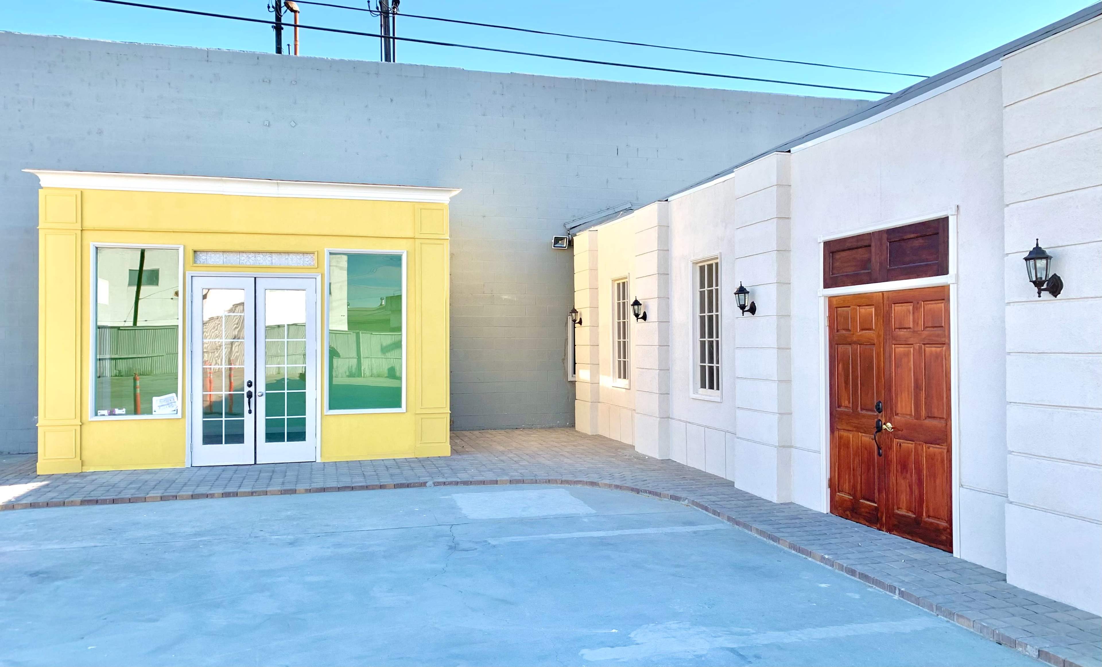 A corner of a building complex with a yellow storefront on the left and a white building with a wooden door on the right.