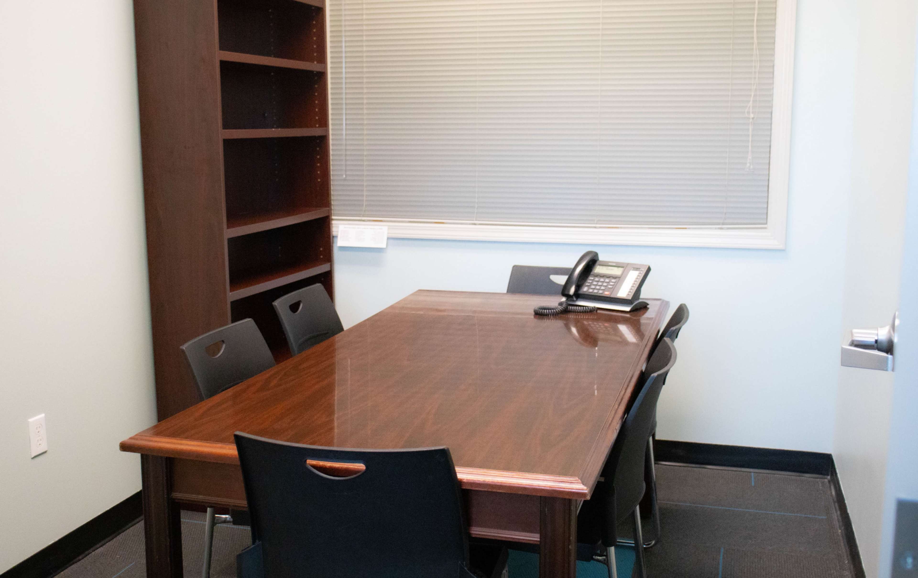 The image shows a small conference room with a wooden table, five black chairs, a telephone, and a bookshelf against the wall.
