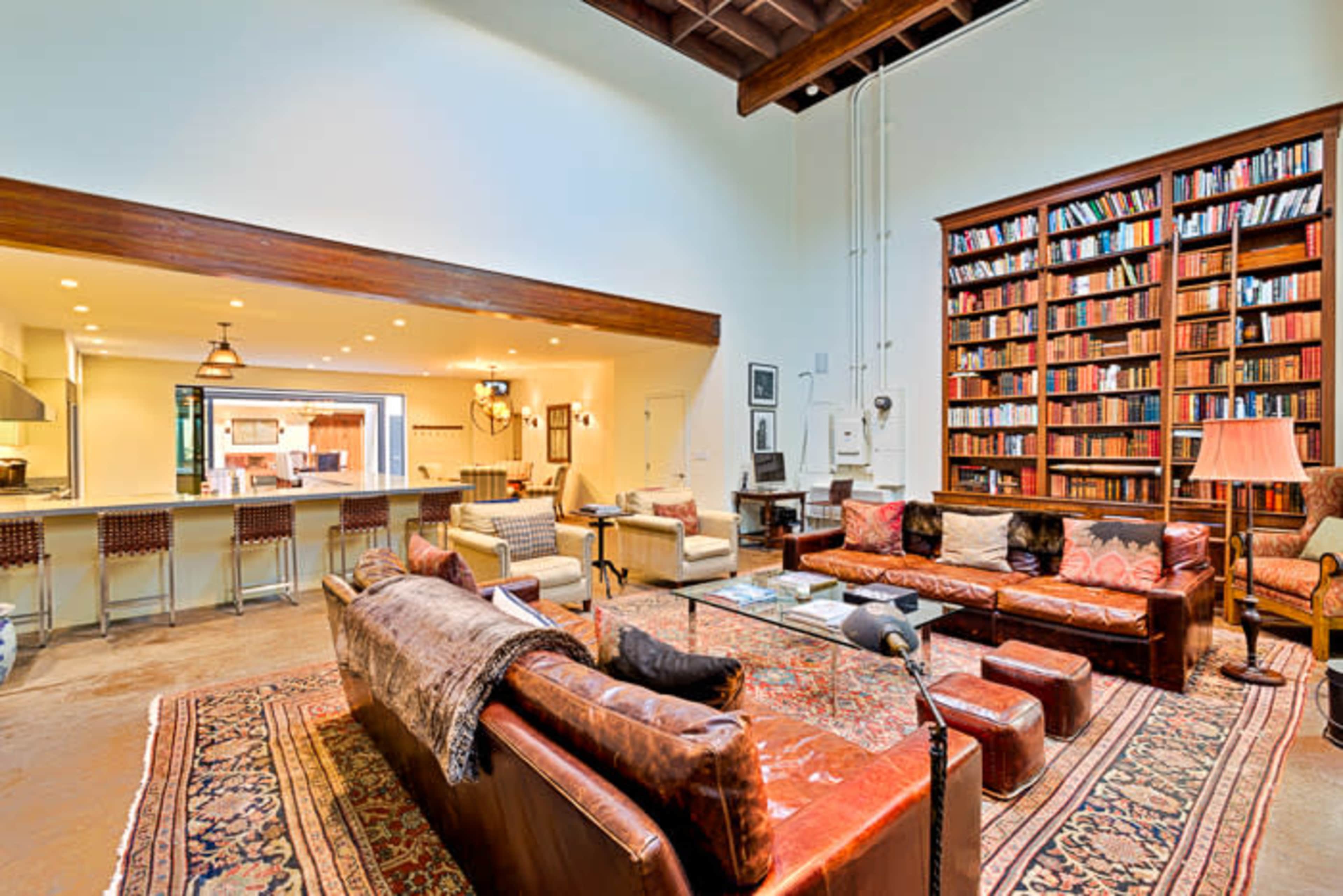 A spacious living room features a large bookshelf, a seating area with leather sofas, and an adjacent kitchen with bar seating.