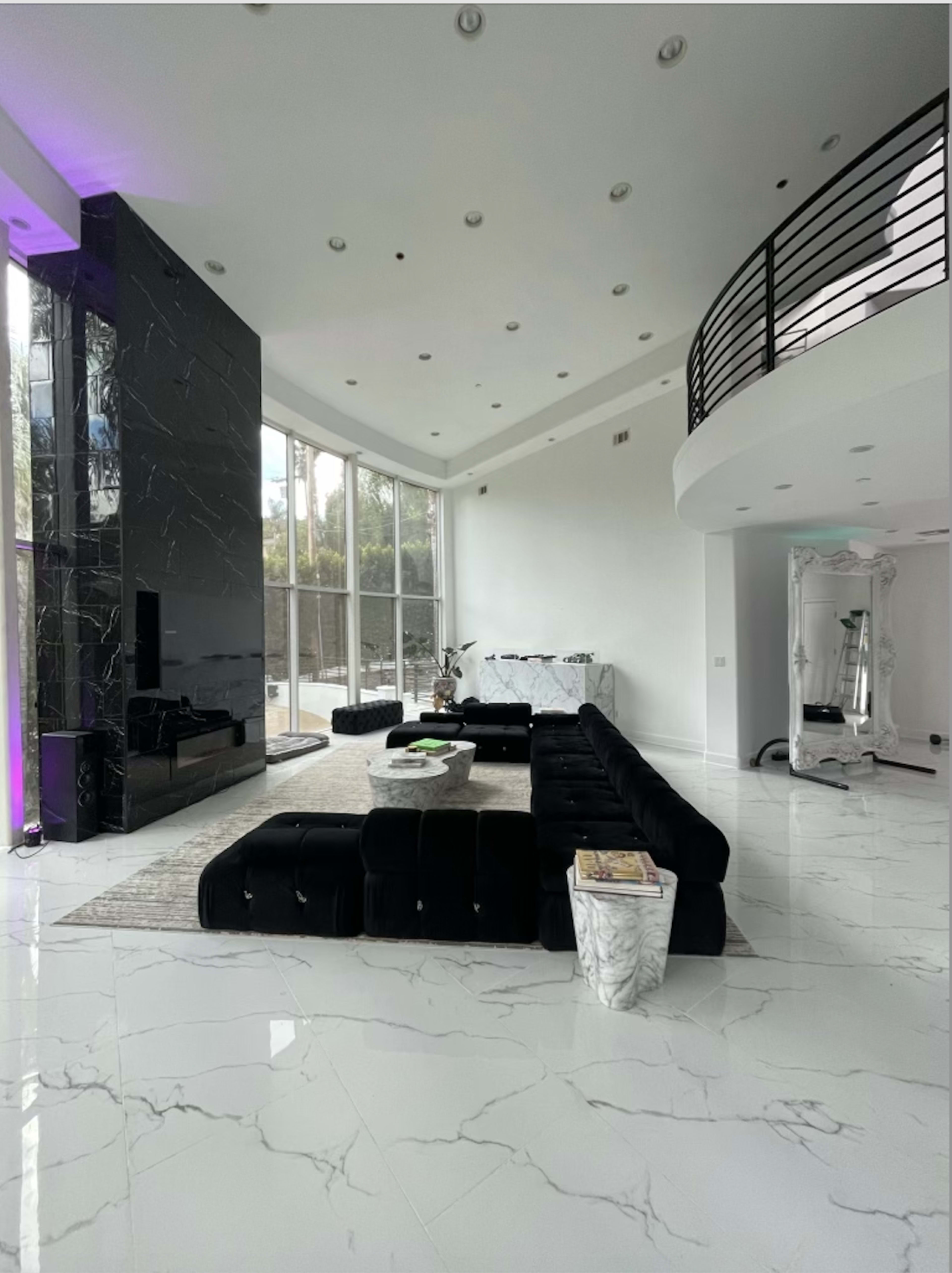 The image shows a modern living room with a large black sectional sofa, a marble floor, and large windows letting in natural light.
