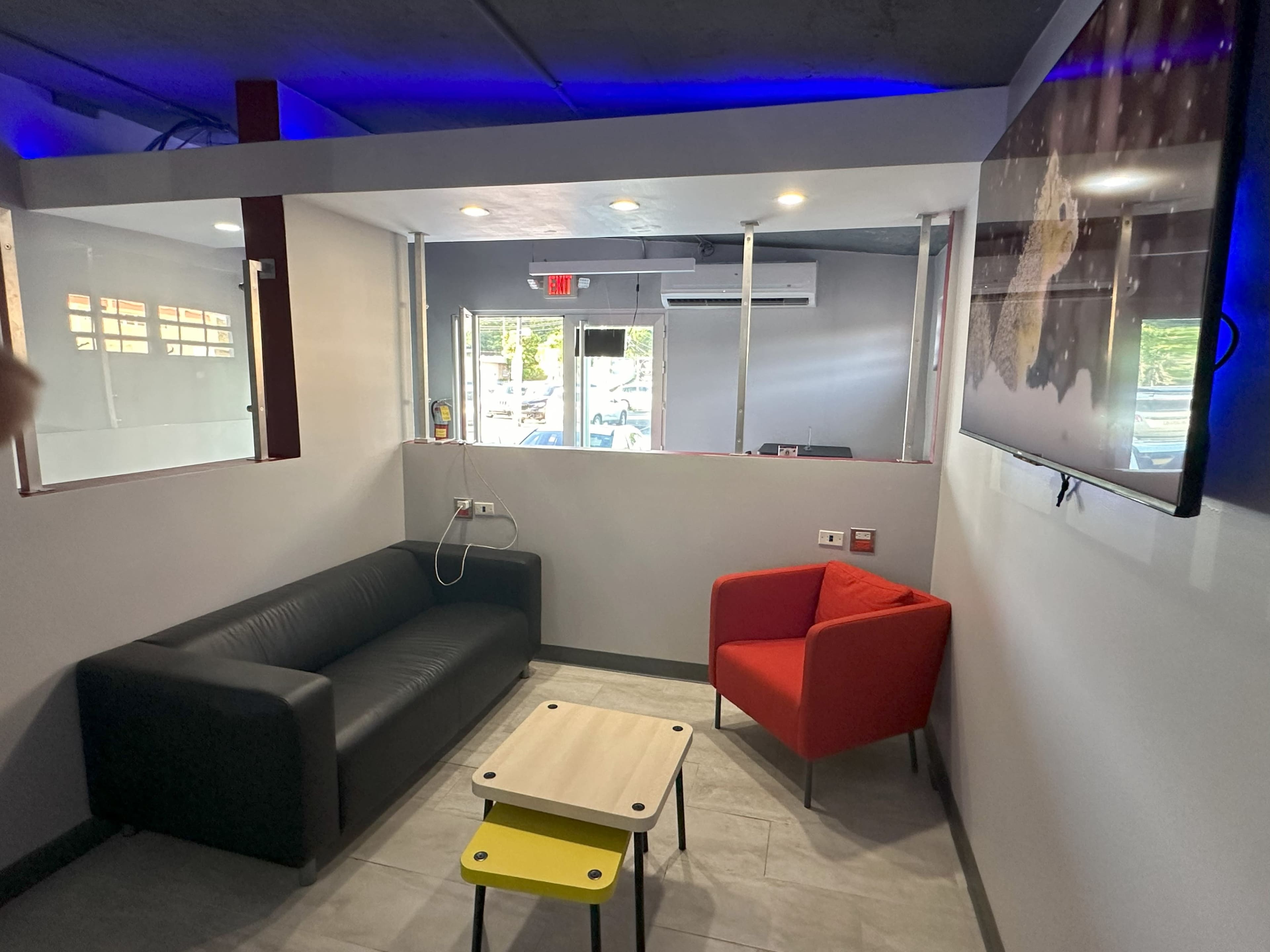 The image shows a small lounge area featuring a black couch, a red armchair, a coffee table, and a wall-mounted television.