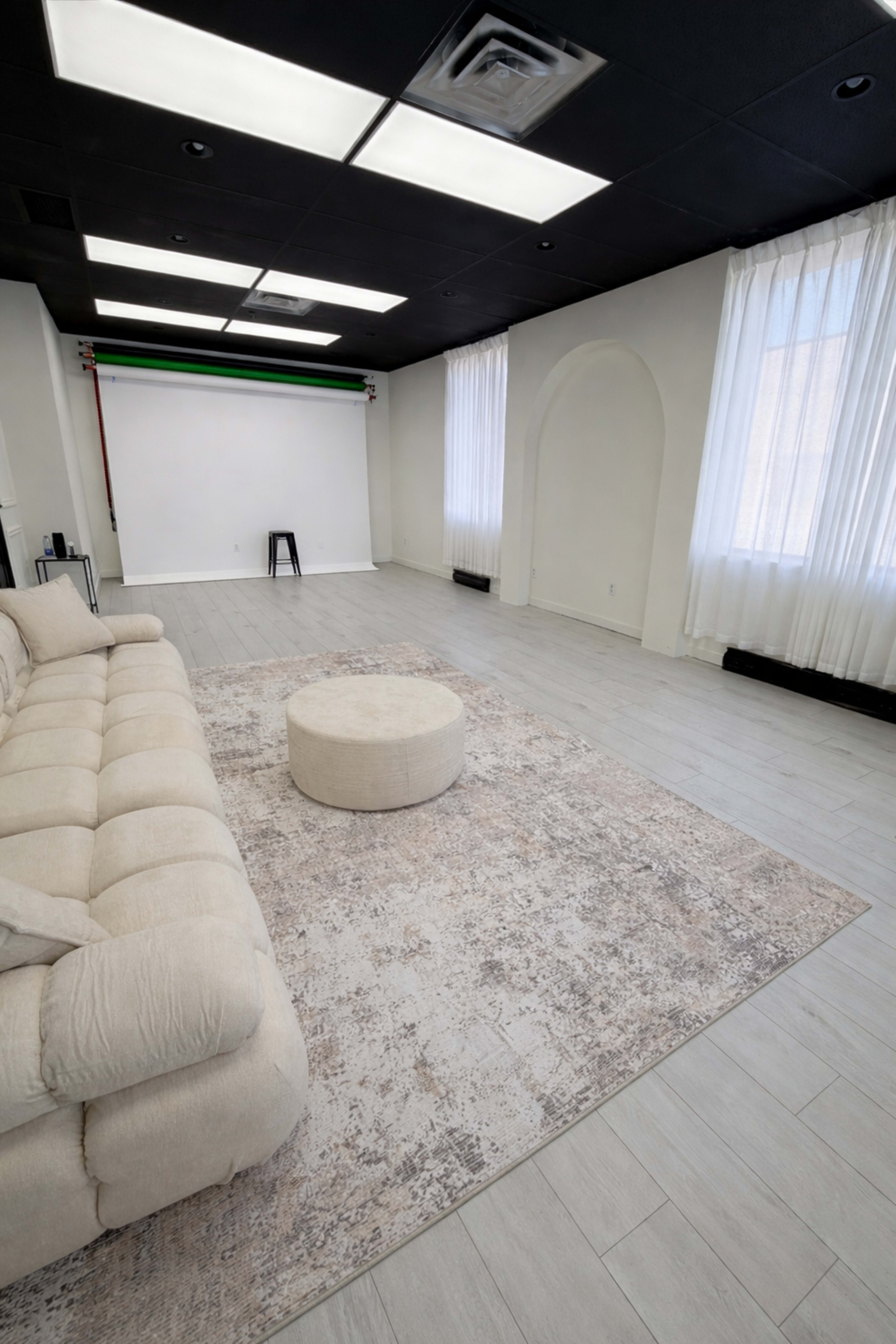 The image shows a spacious indoor studio with a light-colored couch, a round ottoman on a patterned rug, and a backdrop setup for photography behind a black stool.