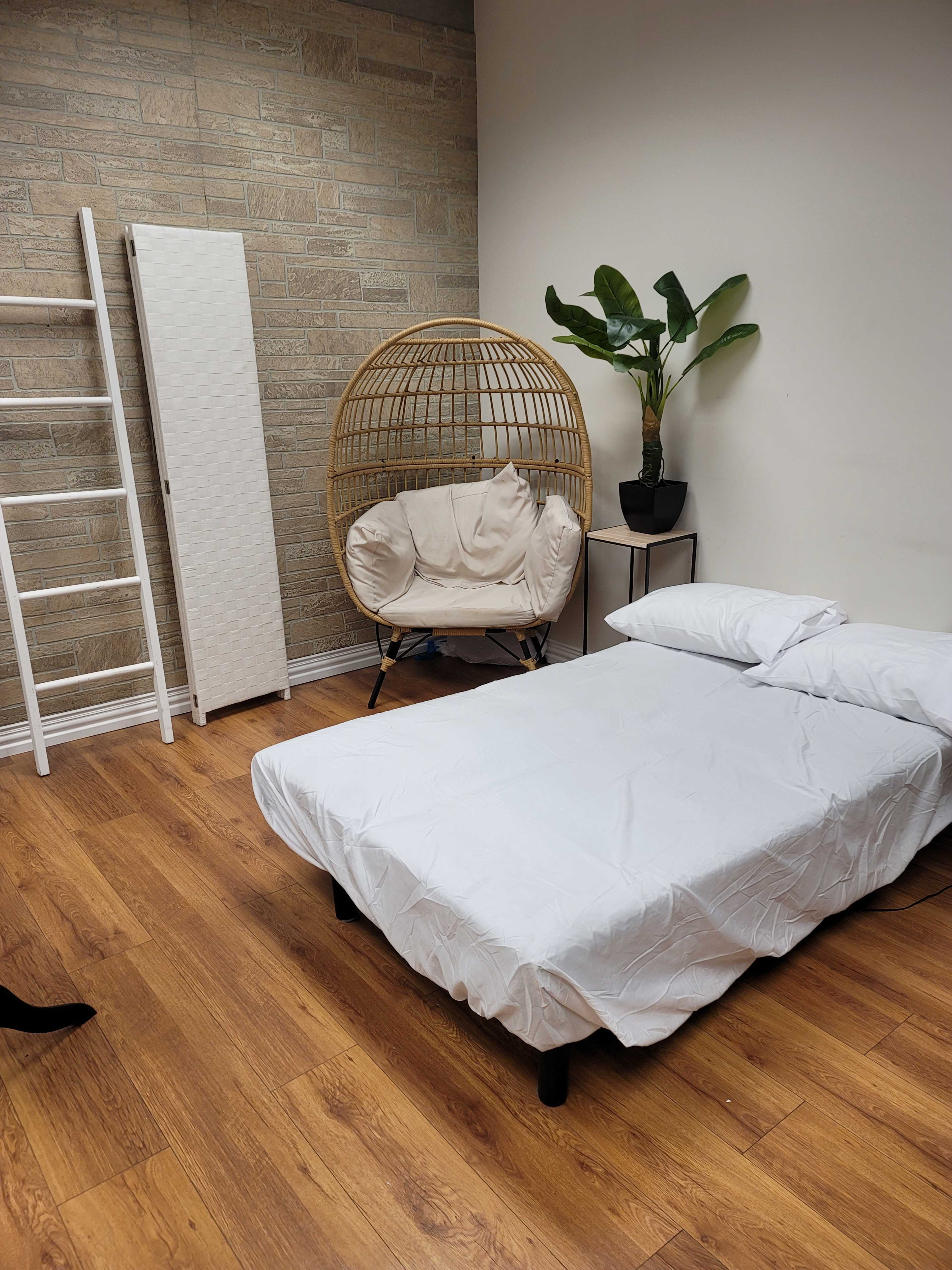 A bedroom with a bed covered in a white sheet, a round wicker chair, a potted plant, and a white folding screen against a textured wall.